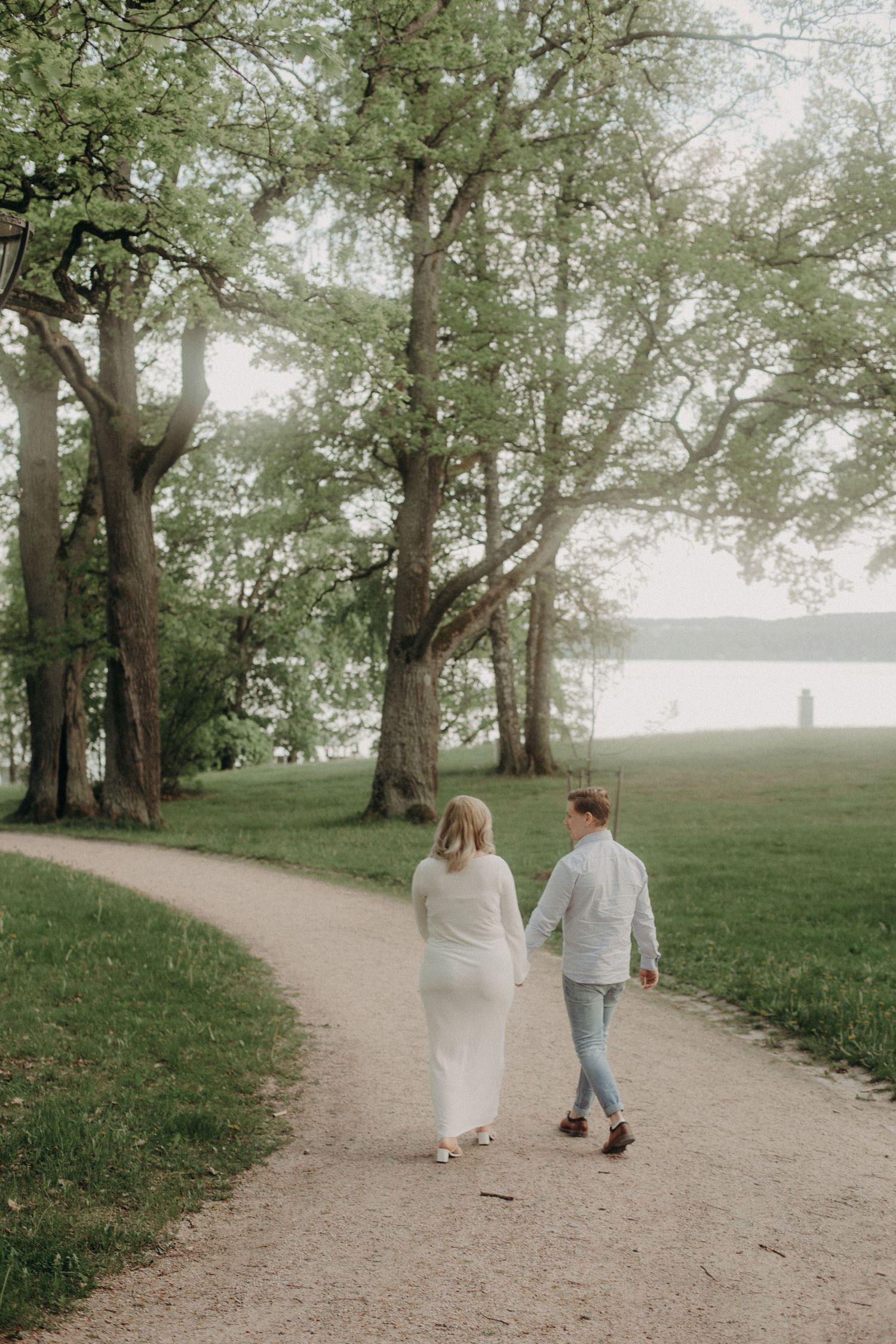 Couple holding hands walking on a winding park path surrounded by lush green trees near a lake.