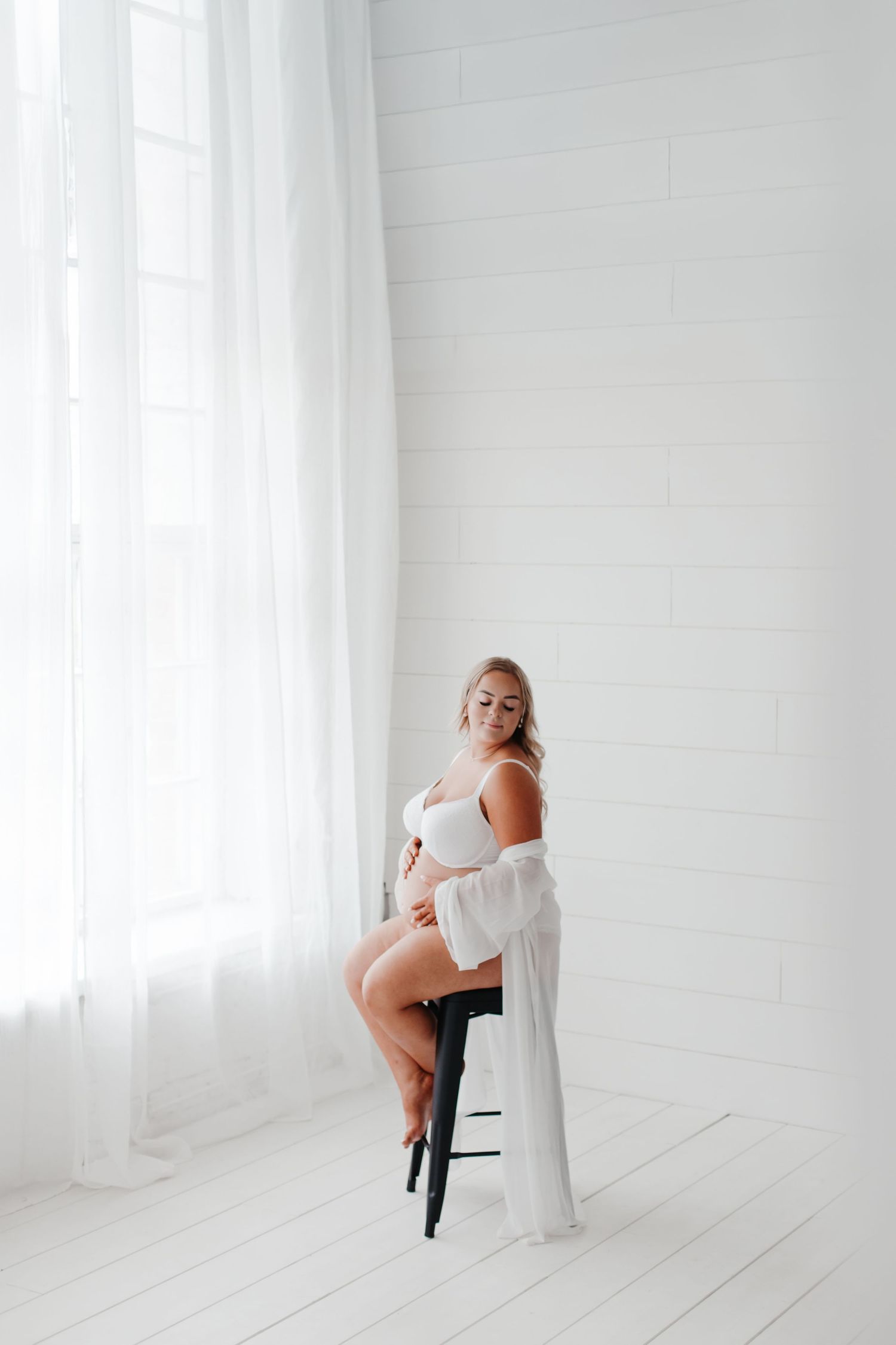 Artistic maternity pose on wooden stool with sheer white robe in bright studio setting.