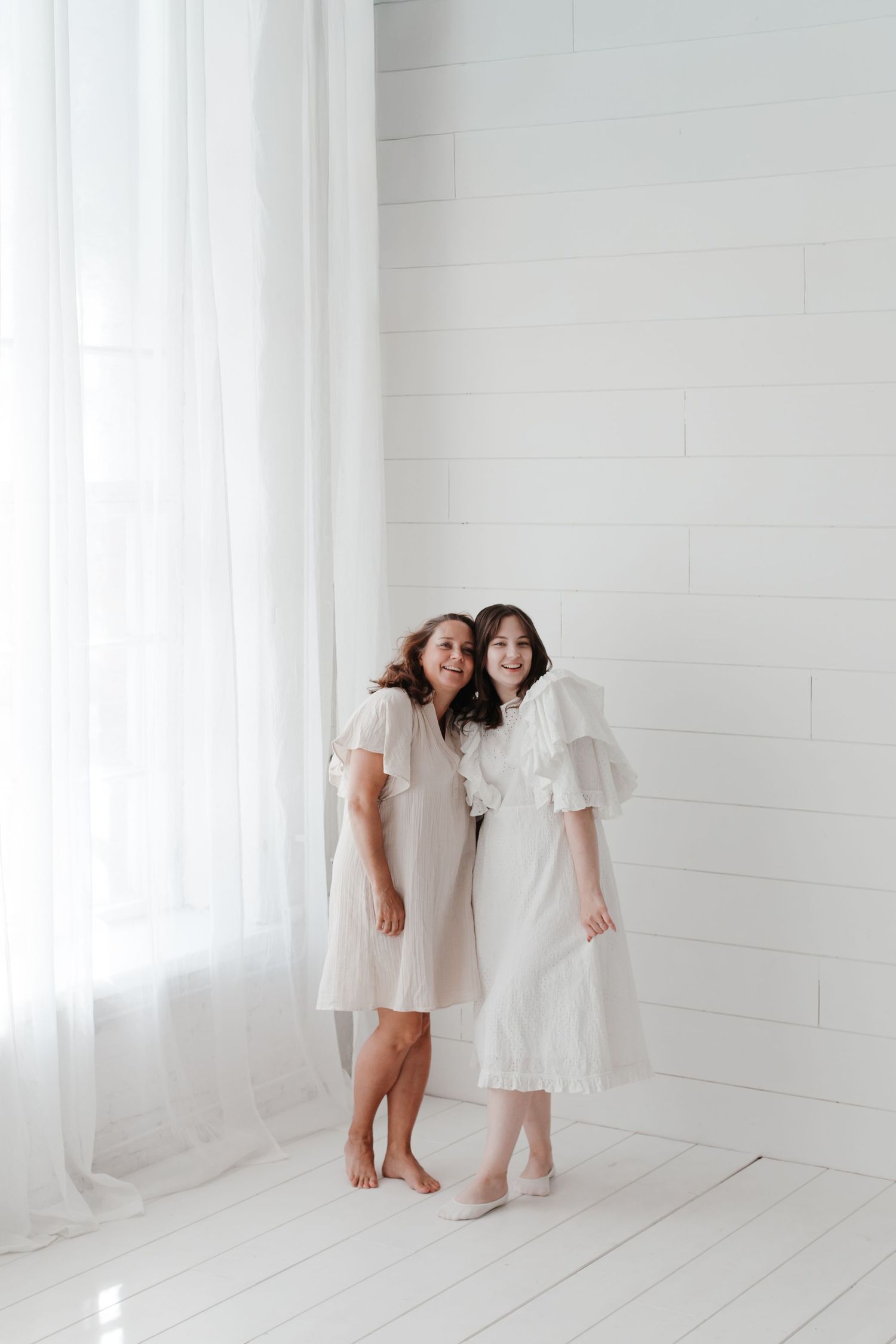 Two people share a joyful moment while wearing white dresses in a bright studio.