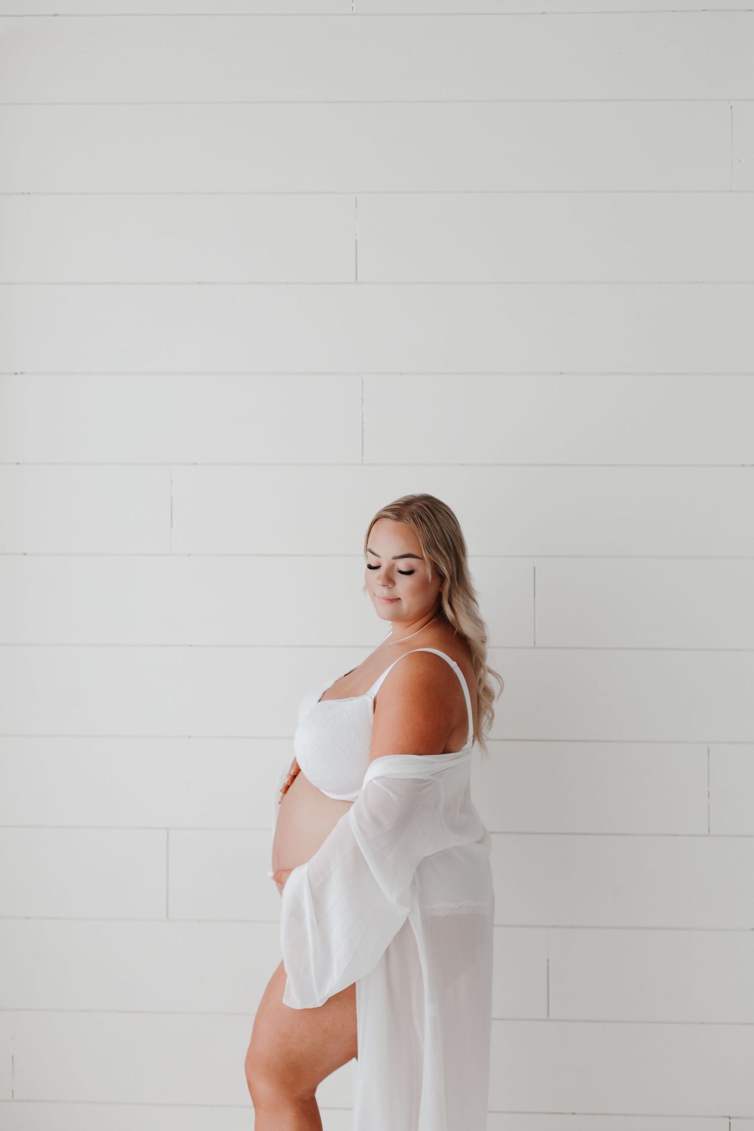 Minimalist maternity photo with white sheer robe against shiplap wall background.