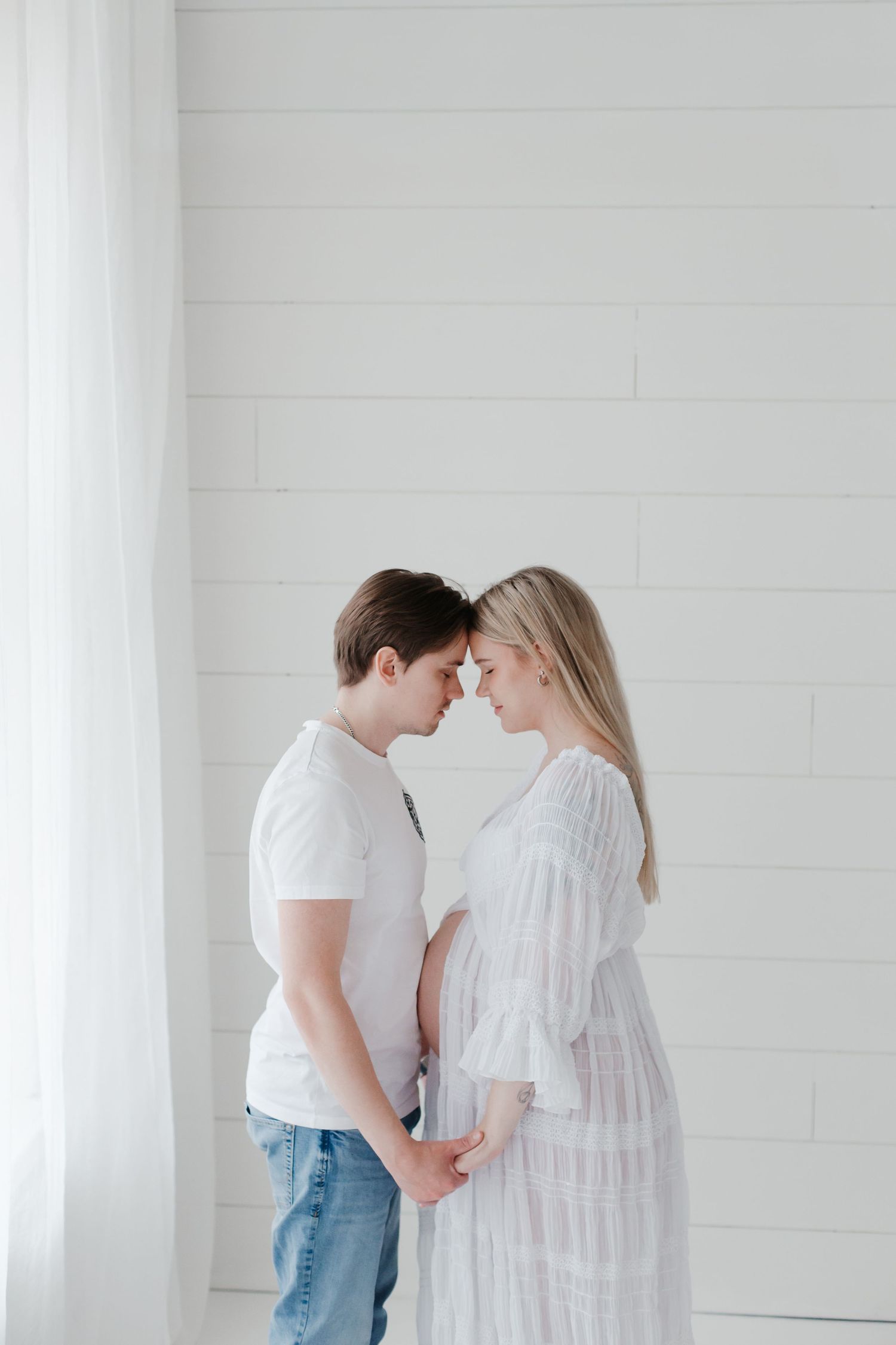 Intimate maternity photoshoot of couple in white against light wall showing tender moment.