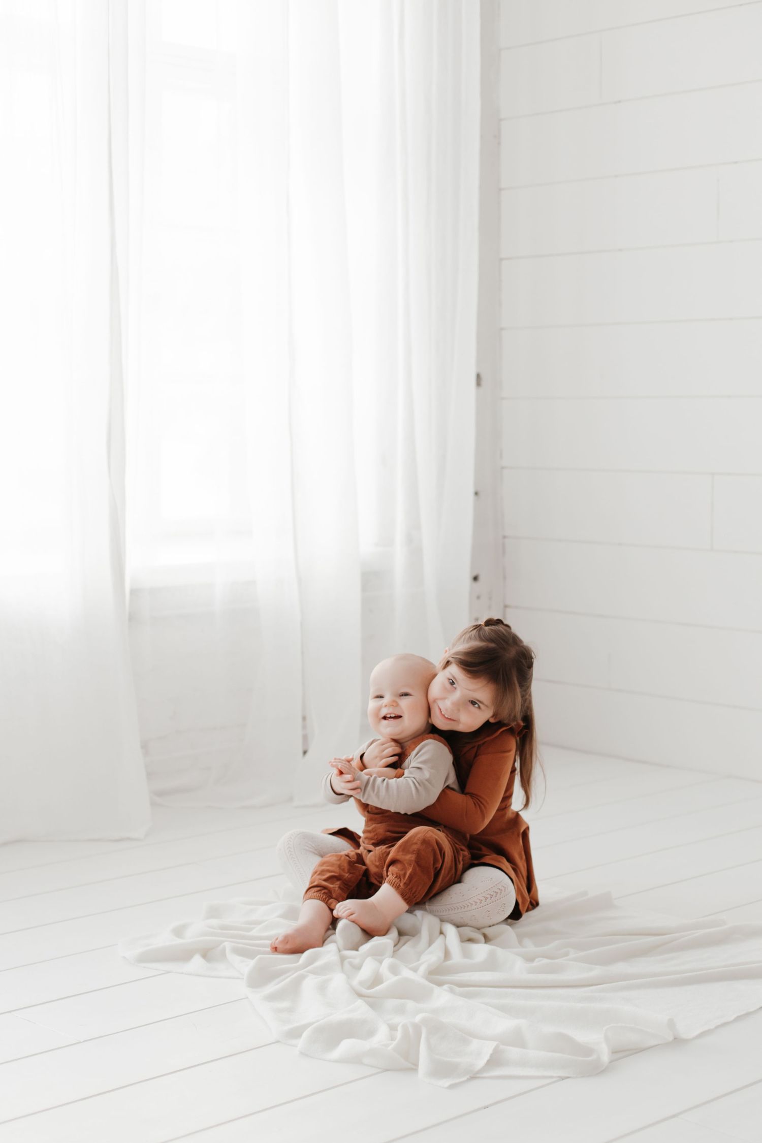 Two children in rust-colored outfits share a sweet moment together on white floor bedding near a window.