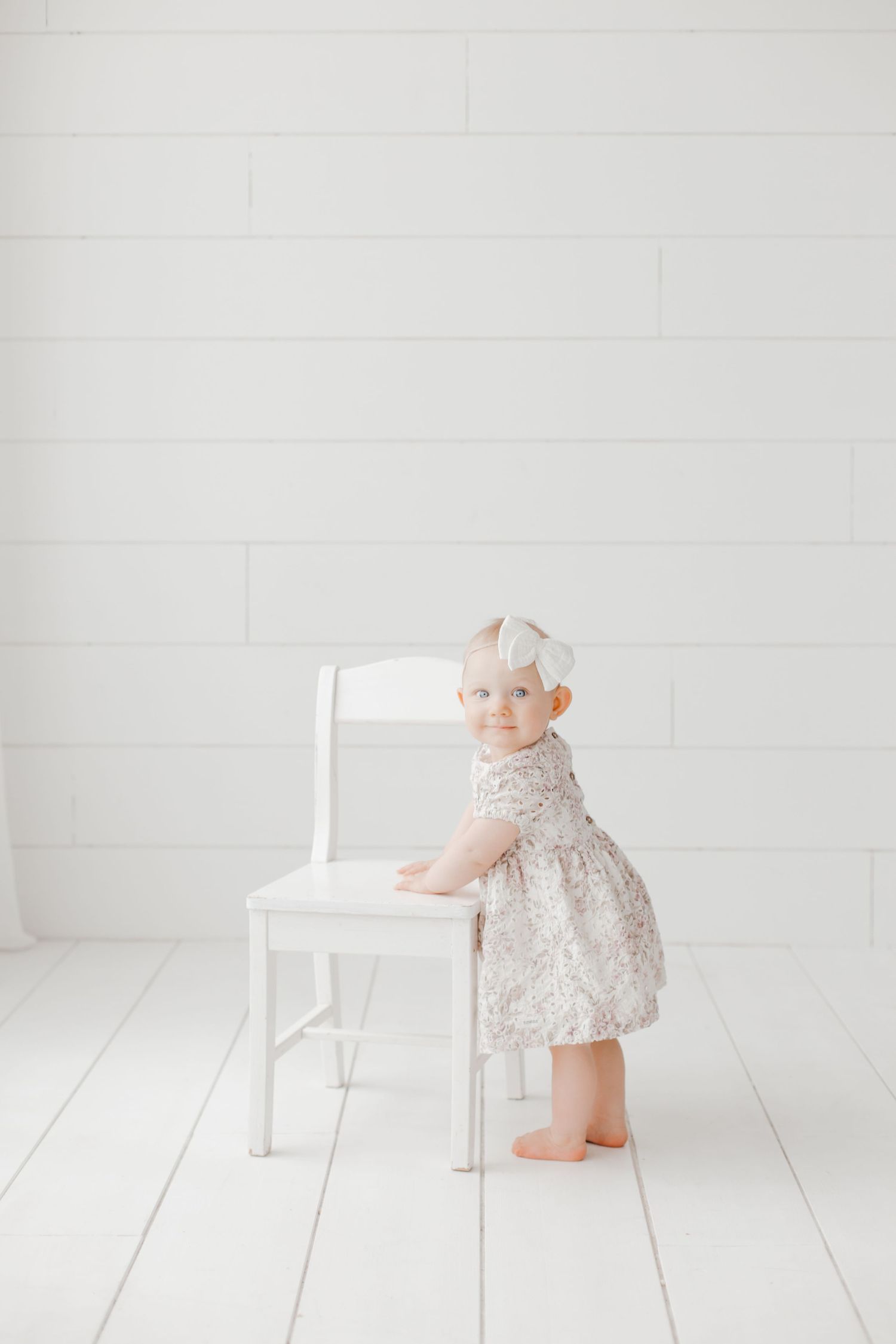 A small child in a lace dress stands next to a white chair in a minimalist white room setting.