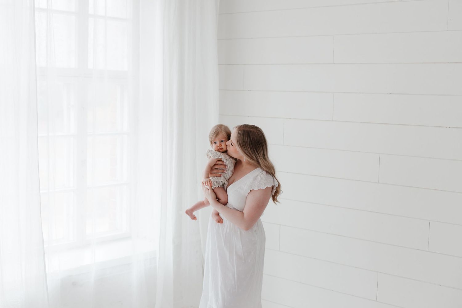 A mother in a white dress holds her baby near a bright window in a minimalist white room.