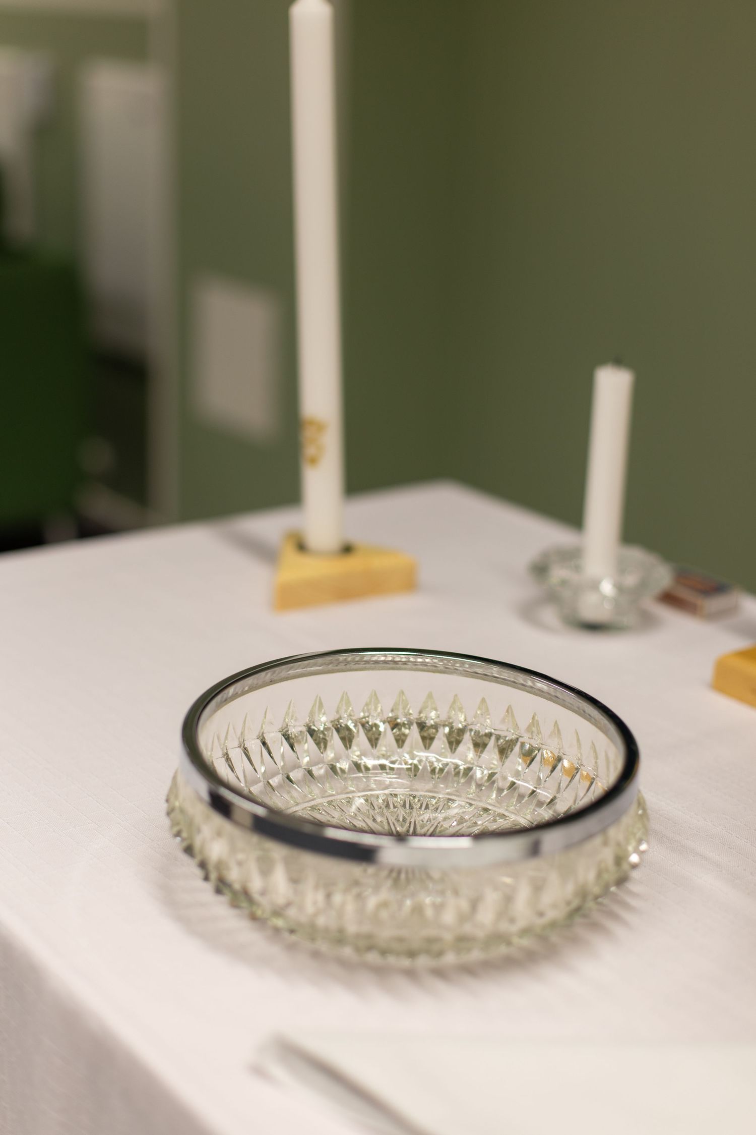 Crystal glass bowl on white tablecloth with white taper candles in gold holders against green wall.