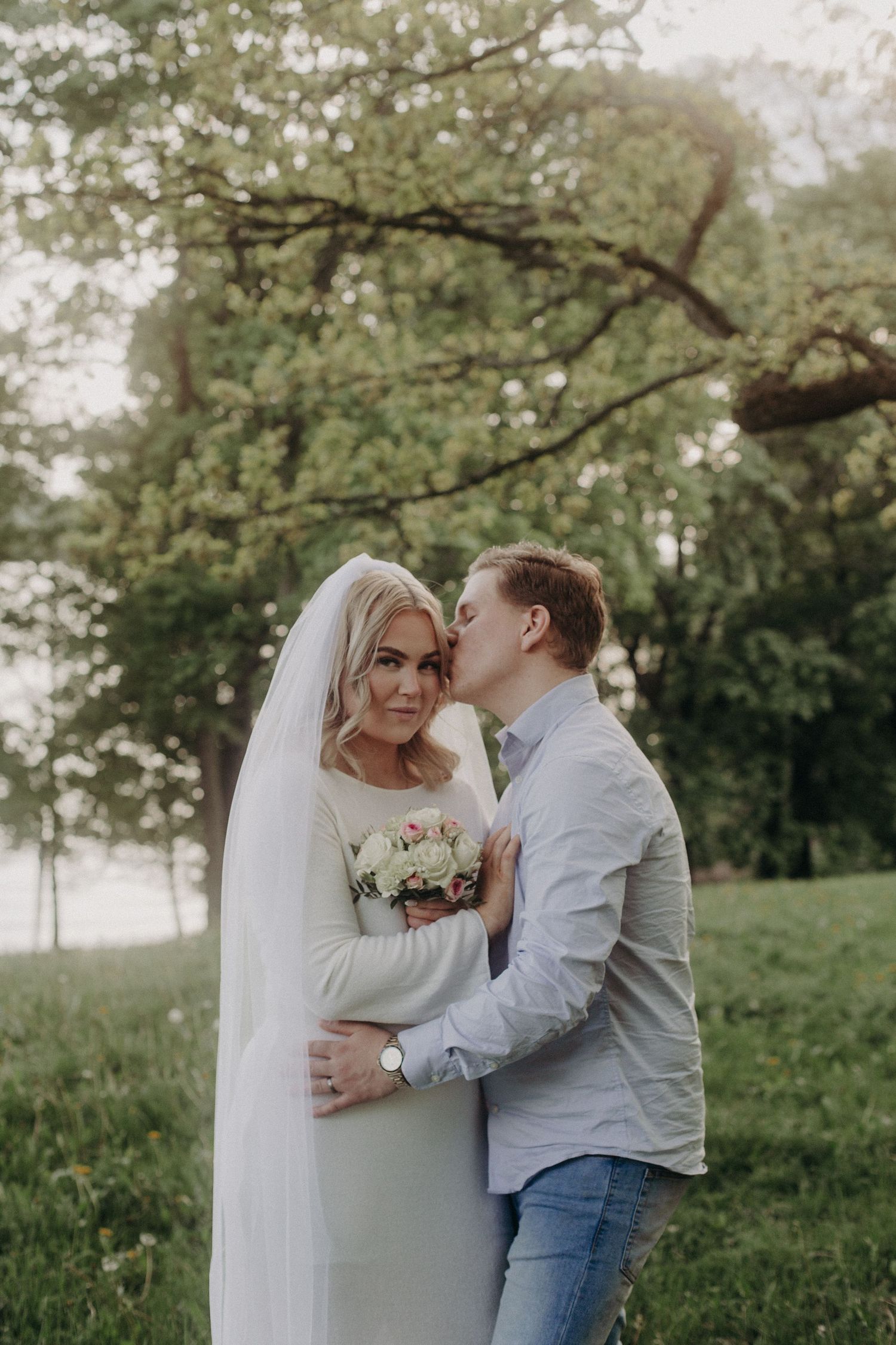 Groom kissing bride on cheek outdoors, bride holding bouquet in white wedding dress and veil.