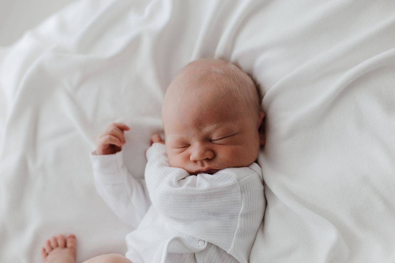 Newborn baby sleeping peacefully in white onesie on soft bedding.