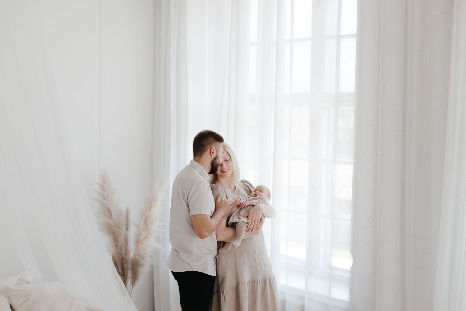 Intimate family portrait by window with soft natural lighting and pampas grass decor.