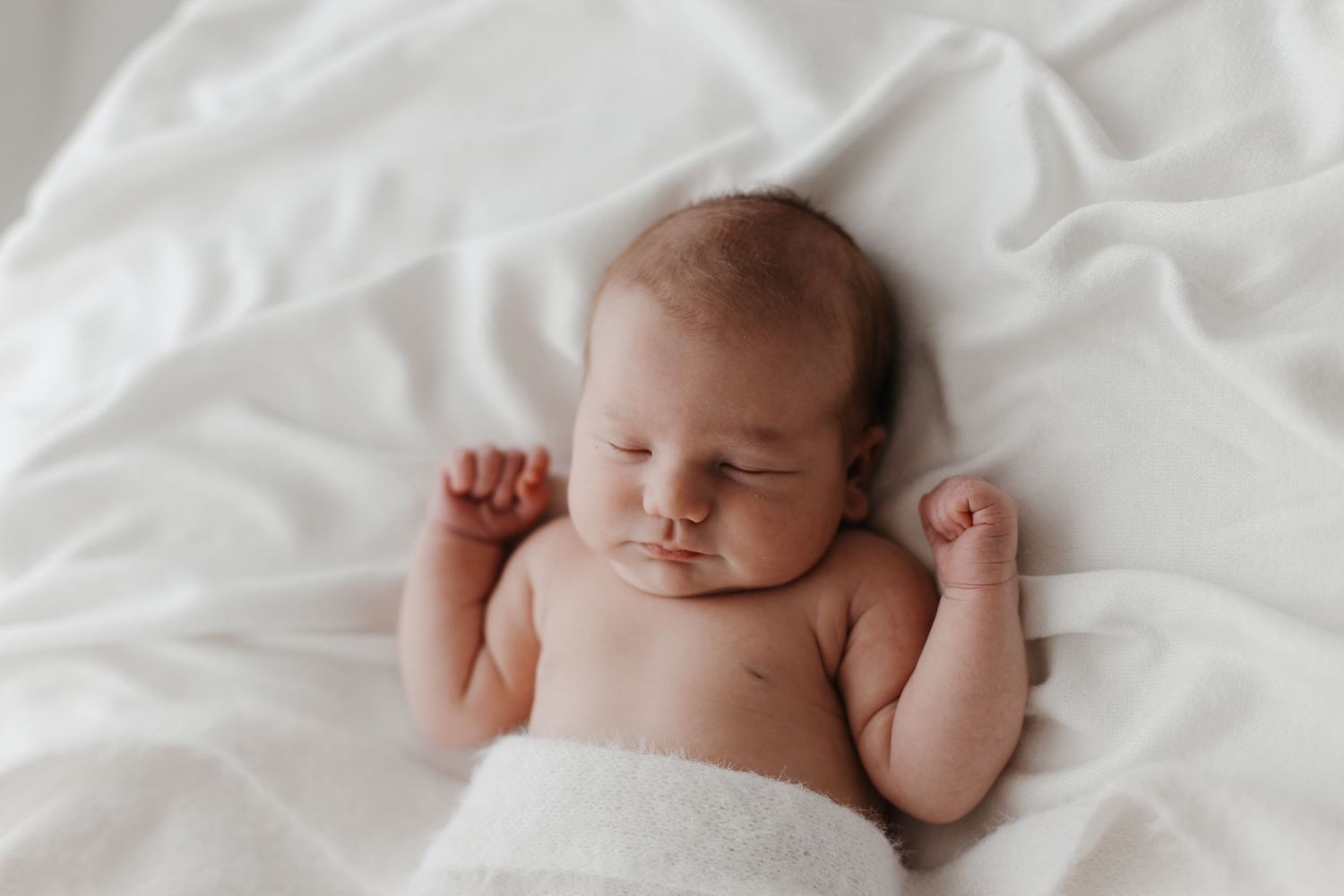 Newborn baby sleeps peacefully on white bedding in natural light.