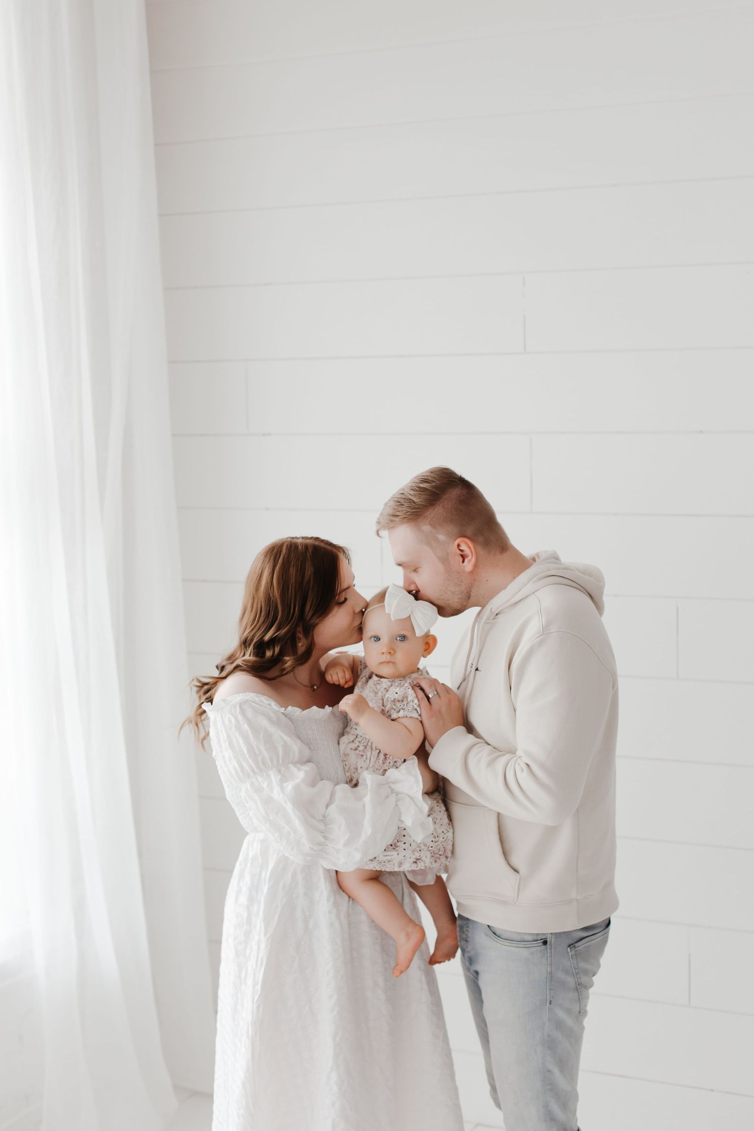 Family wearing white outfits shares tender moments in bright minimalist studio setting.