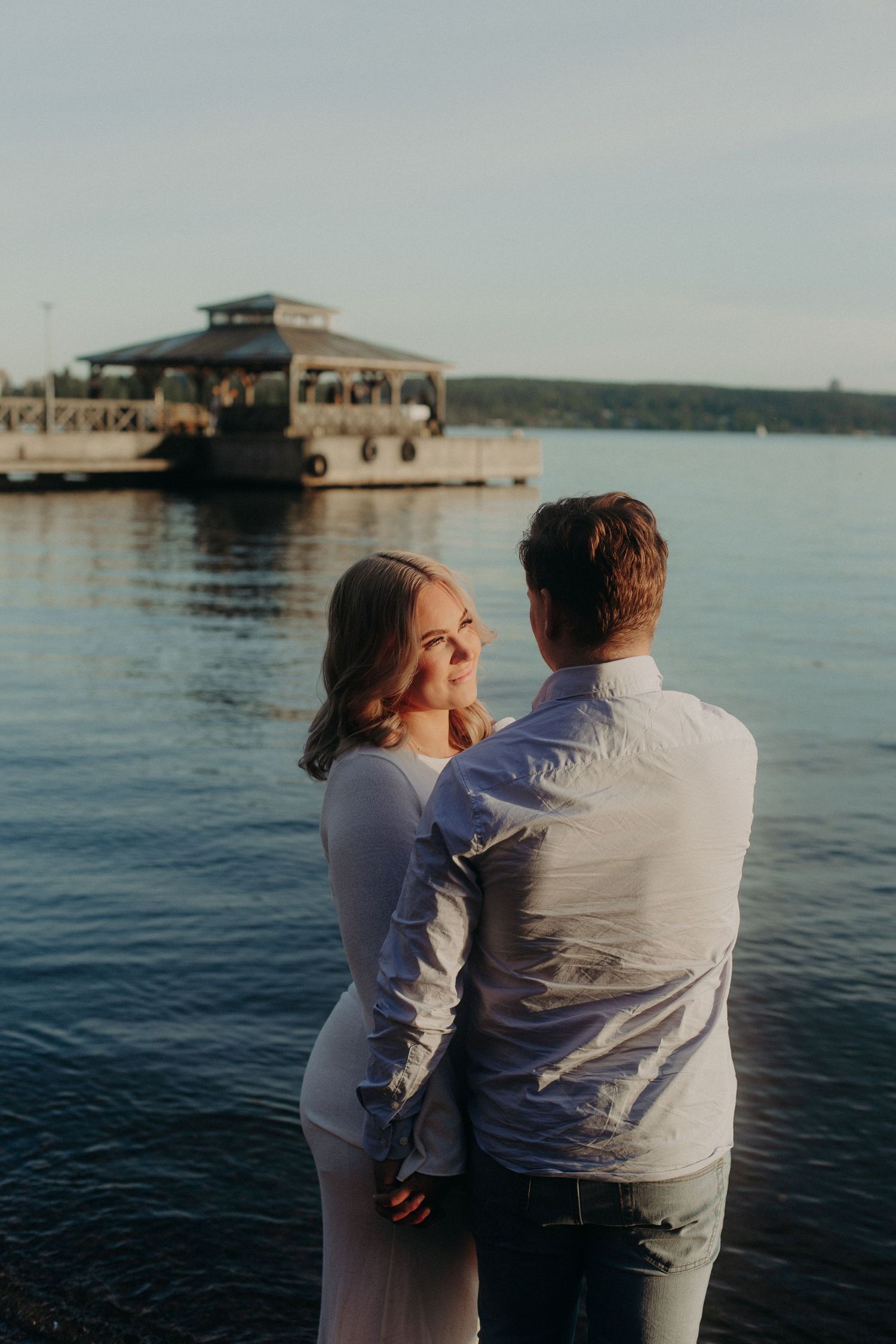 Couple embracing by a lake at sunset, woman in white dress smiling over her shoulder.