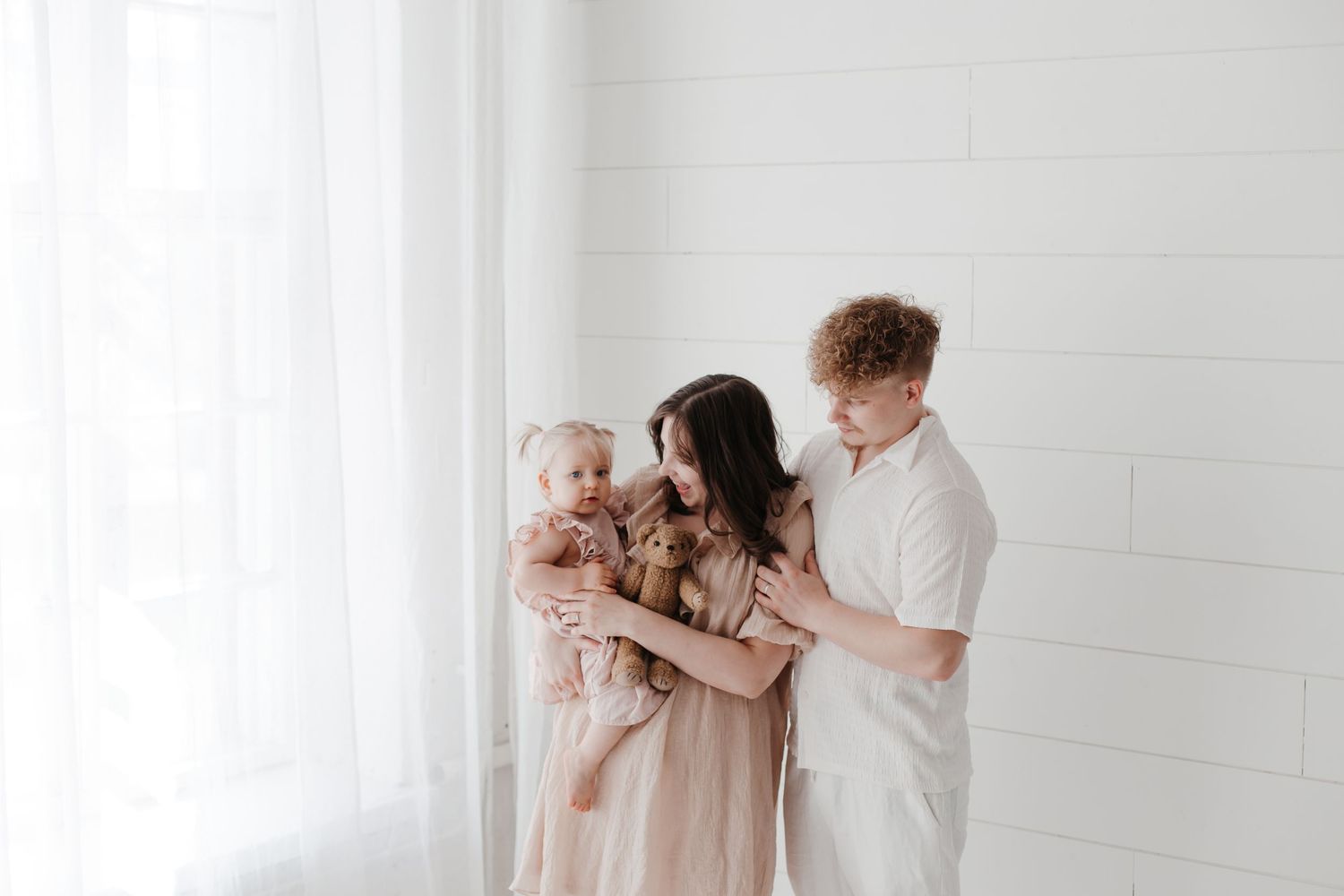 A young family with a redhead father and brunette mother holding their baby girl in a bright white studio.