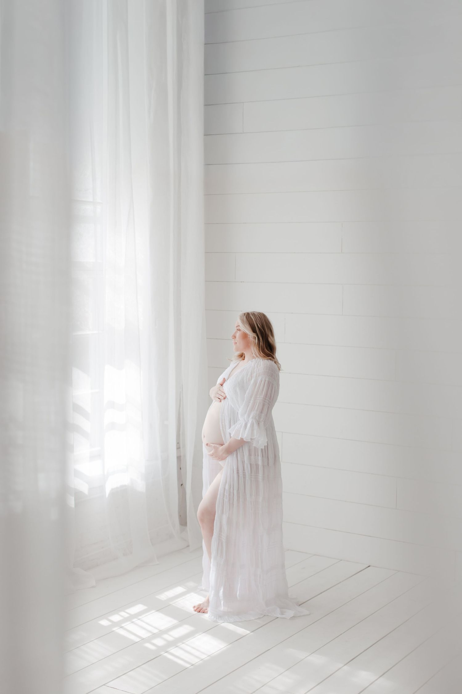 Graceful maternity silhouette in white lace against bright window light.