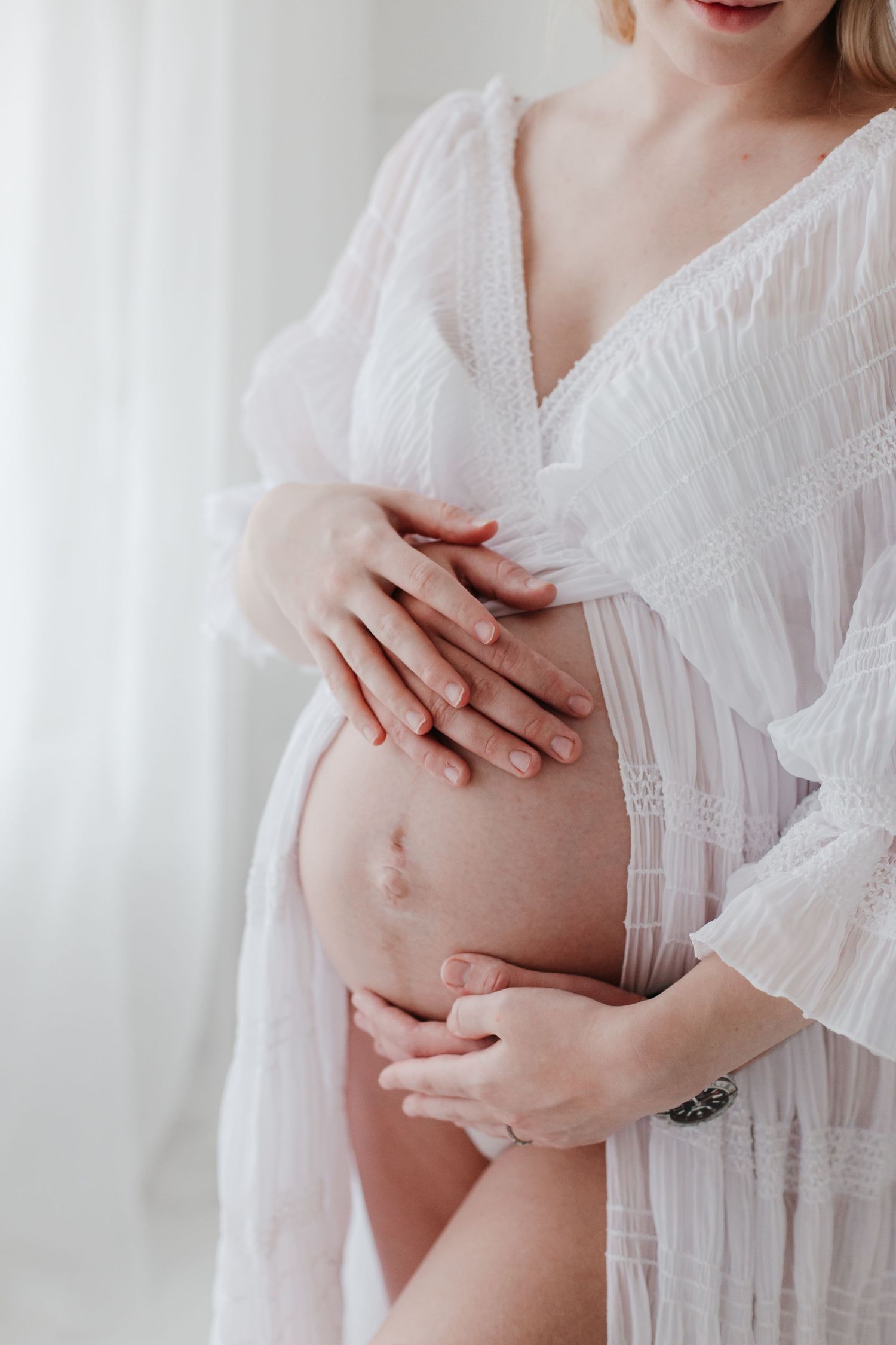 Close-up of pregnant belly with hands gently resting in white lace dress.