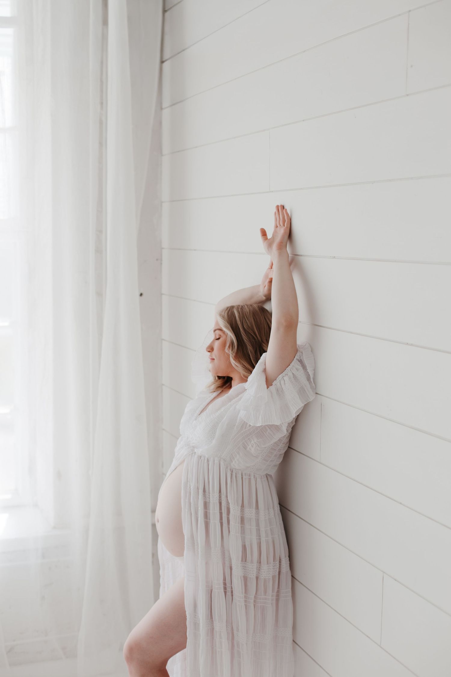 Artistic maternity pose against white shiplap wall in delicate lace gown.