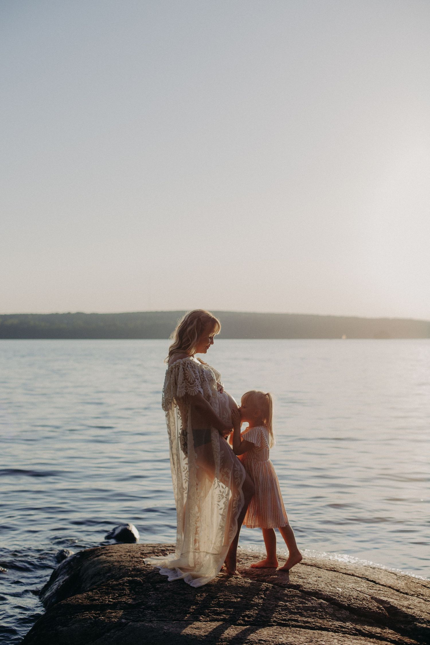 Romantic maternity pose on lakeside rocks with sheer lace dress at dusk.