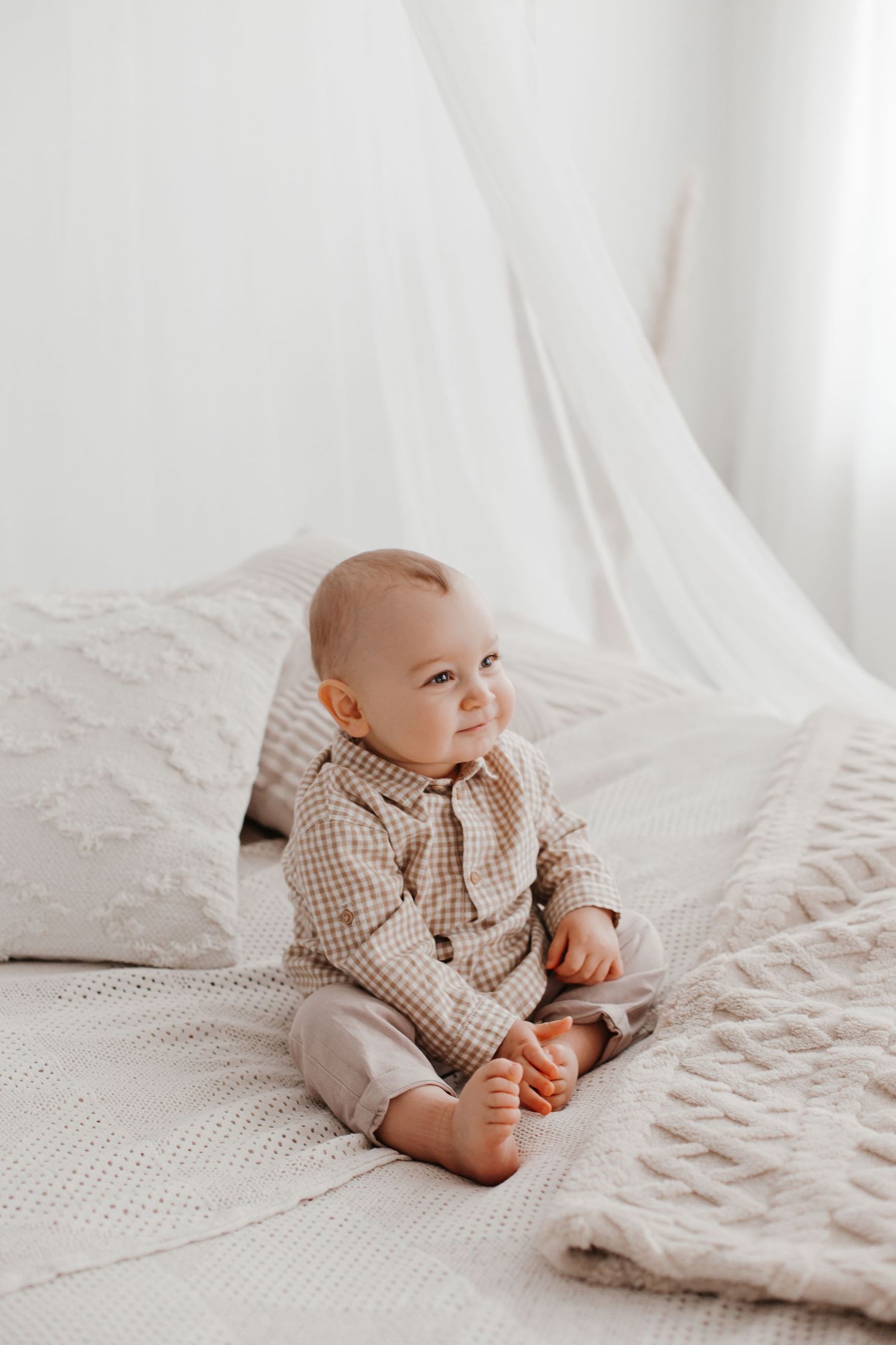 Smiling baby in gingham shirt sitting on white textured bedding in bright room.