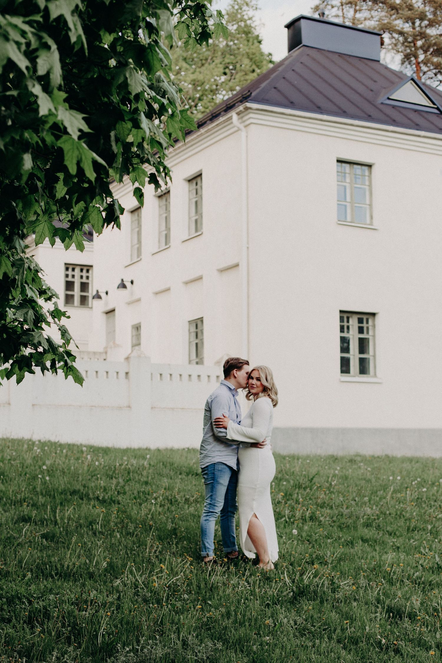 Couple embracing on green lawn before white manor house, sharing romantic moment under leafy tree in summer.