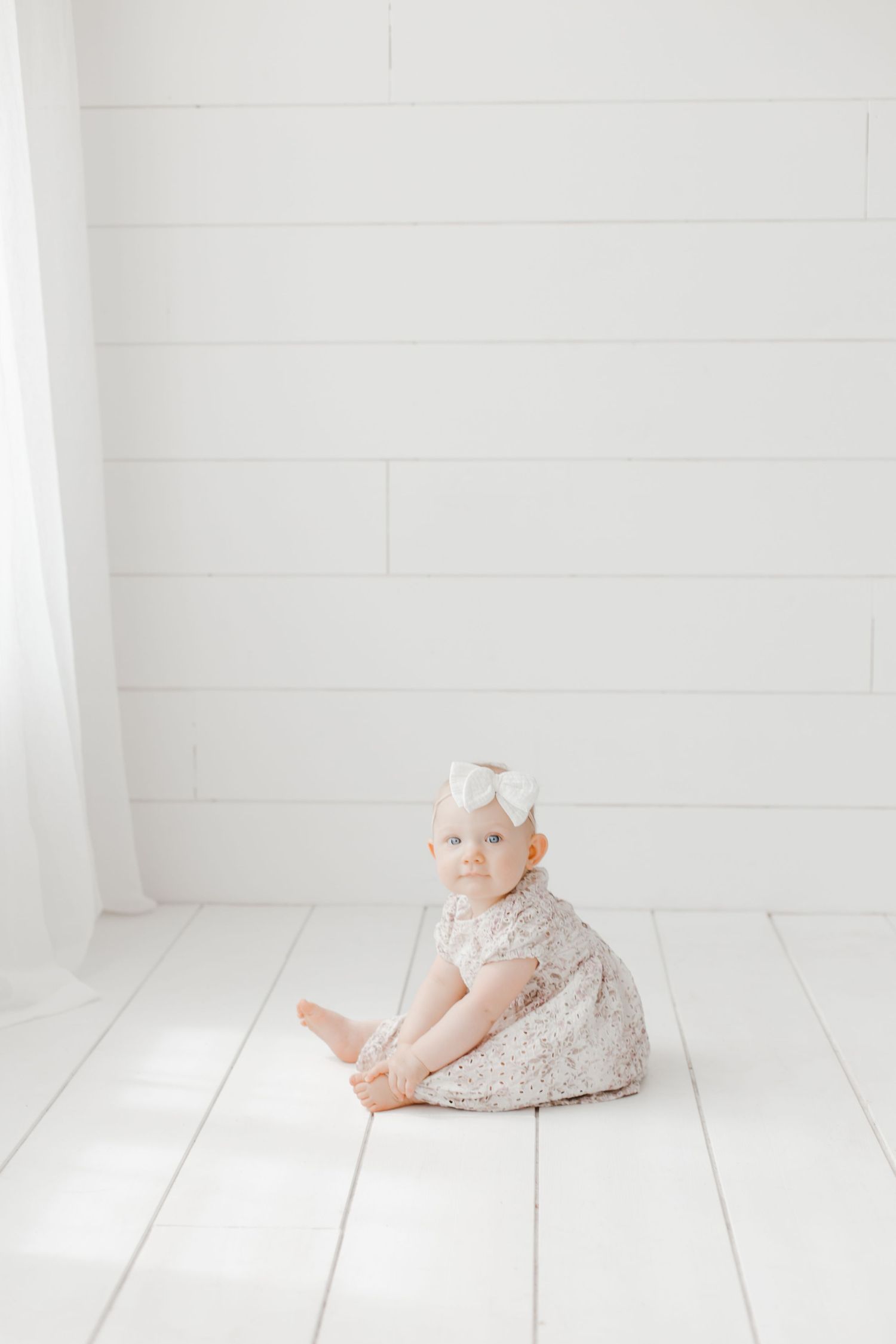 A small child in a lace dress sits on white wooden flooring in a bright, minimal setting.