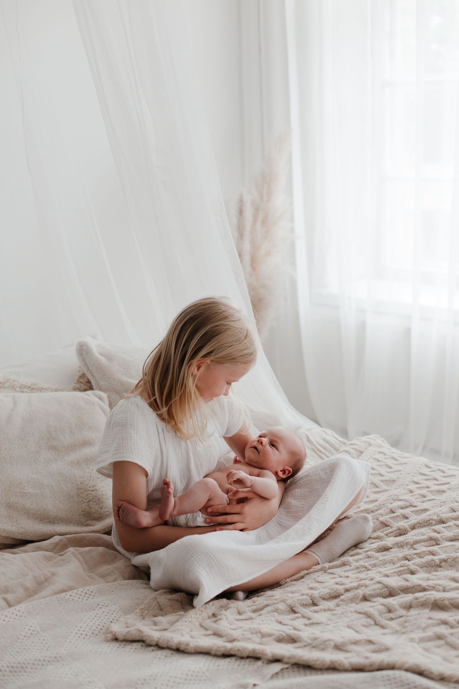 Child in white dress bonding with newborn sibling on textured beige bedding.