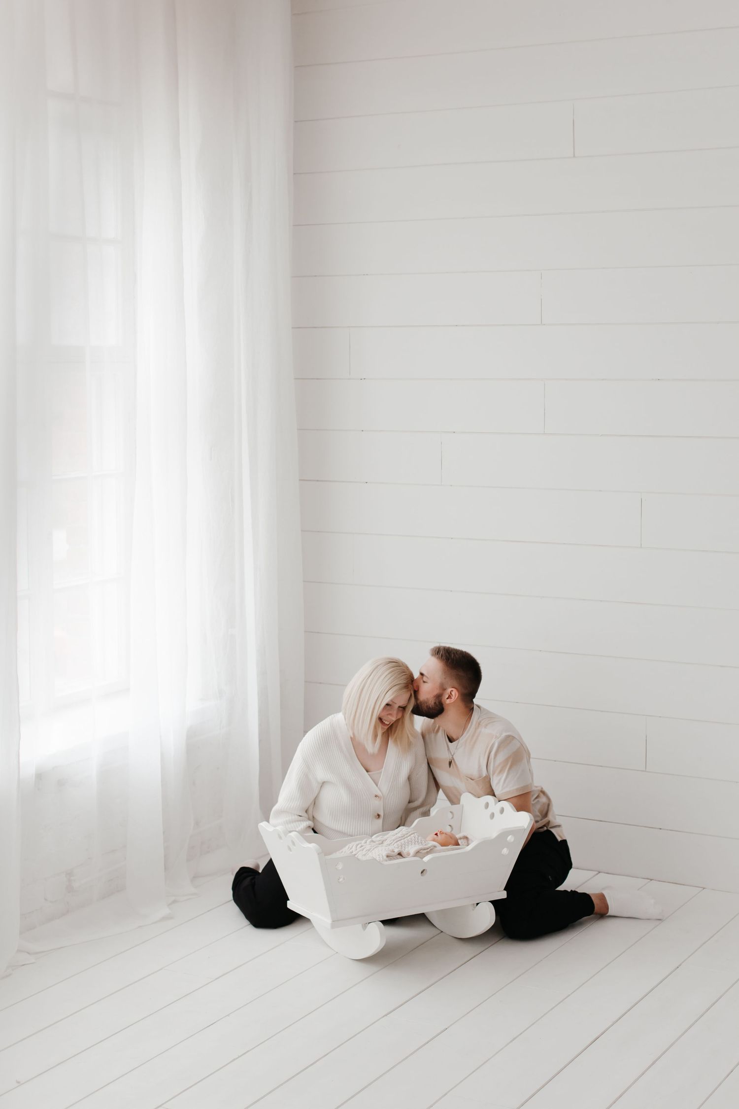 Family in cream sweaters shares tender moment on white wooden floor.