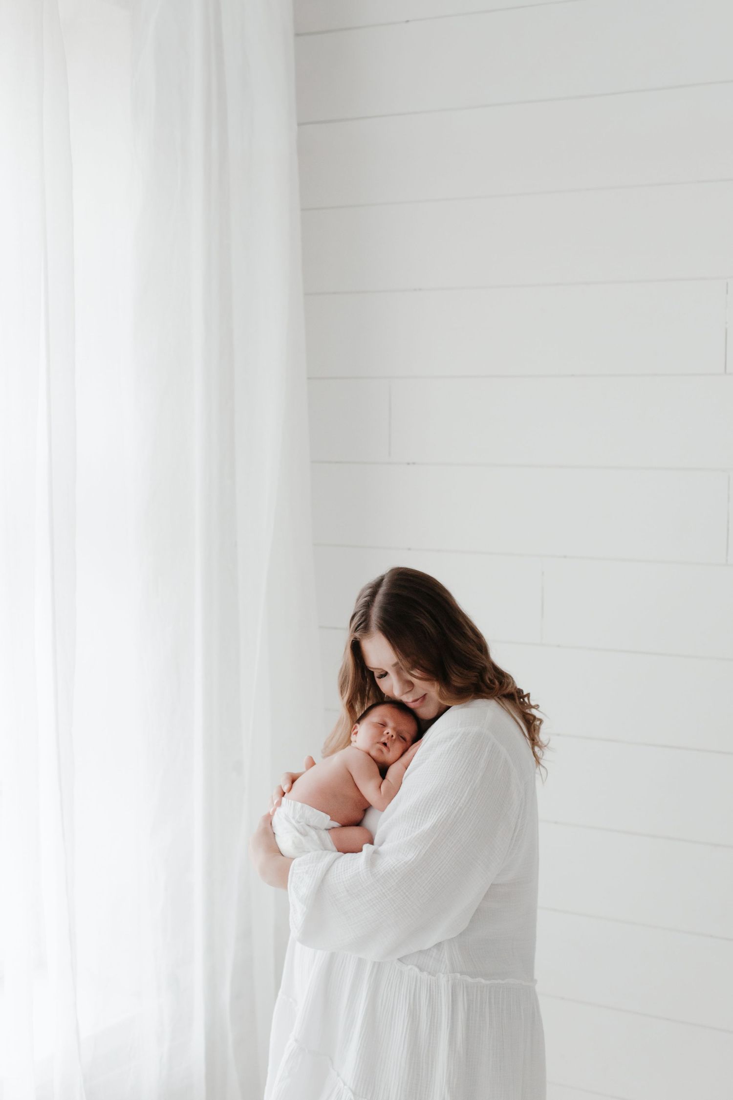 A tender moment captured in white as a mother embraces her newborn baby against a bright minimalist backdrop.
