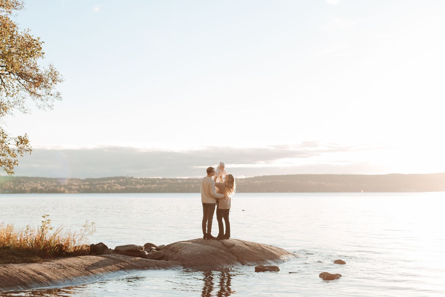 Couple stands on rocky coastline during golden sunset over tranquil waters.