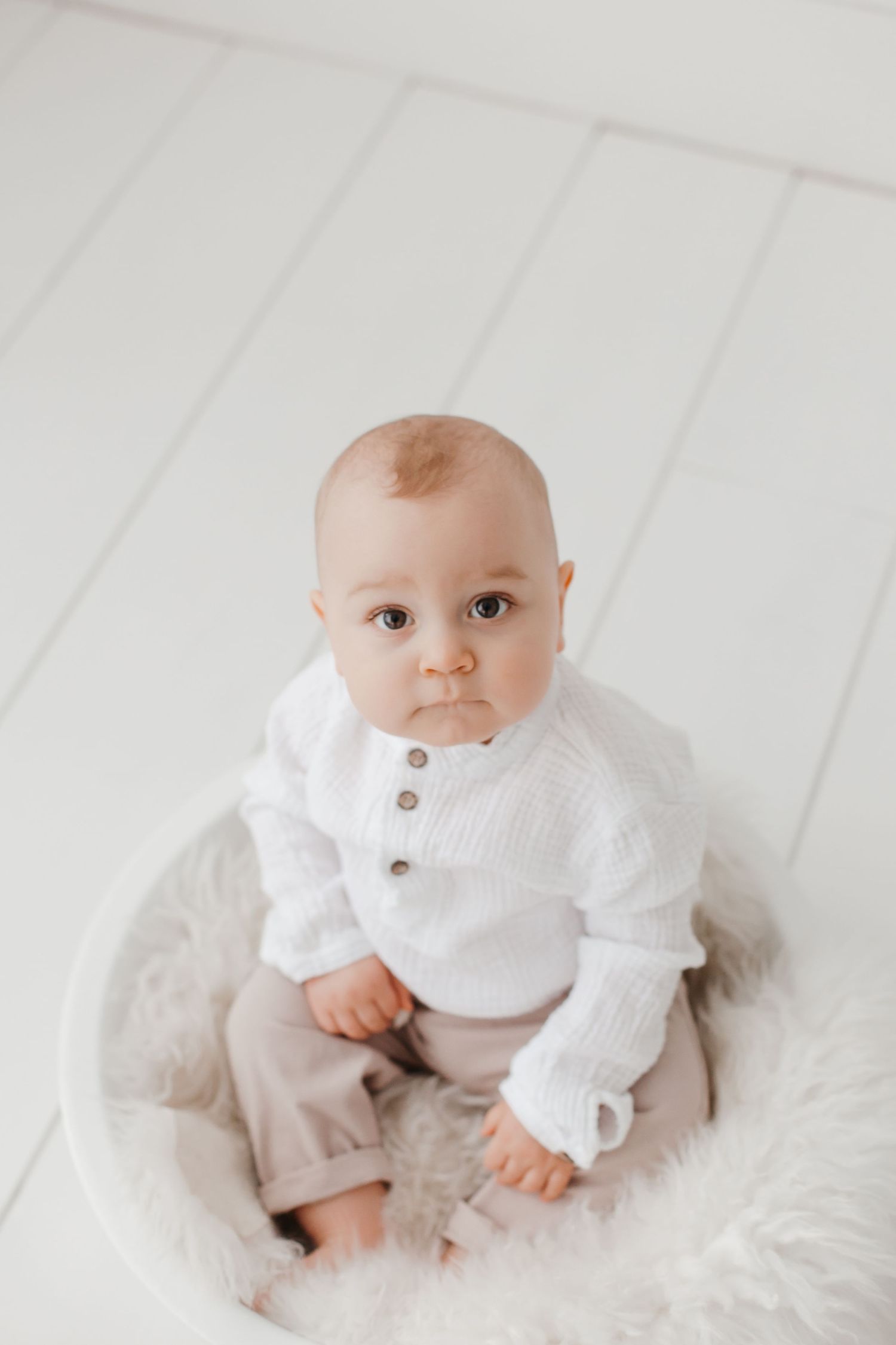 Baby in white sweater and beige pants sitting on fluffy white cushion against white backdrop.
