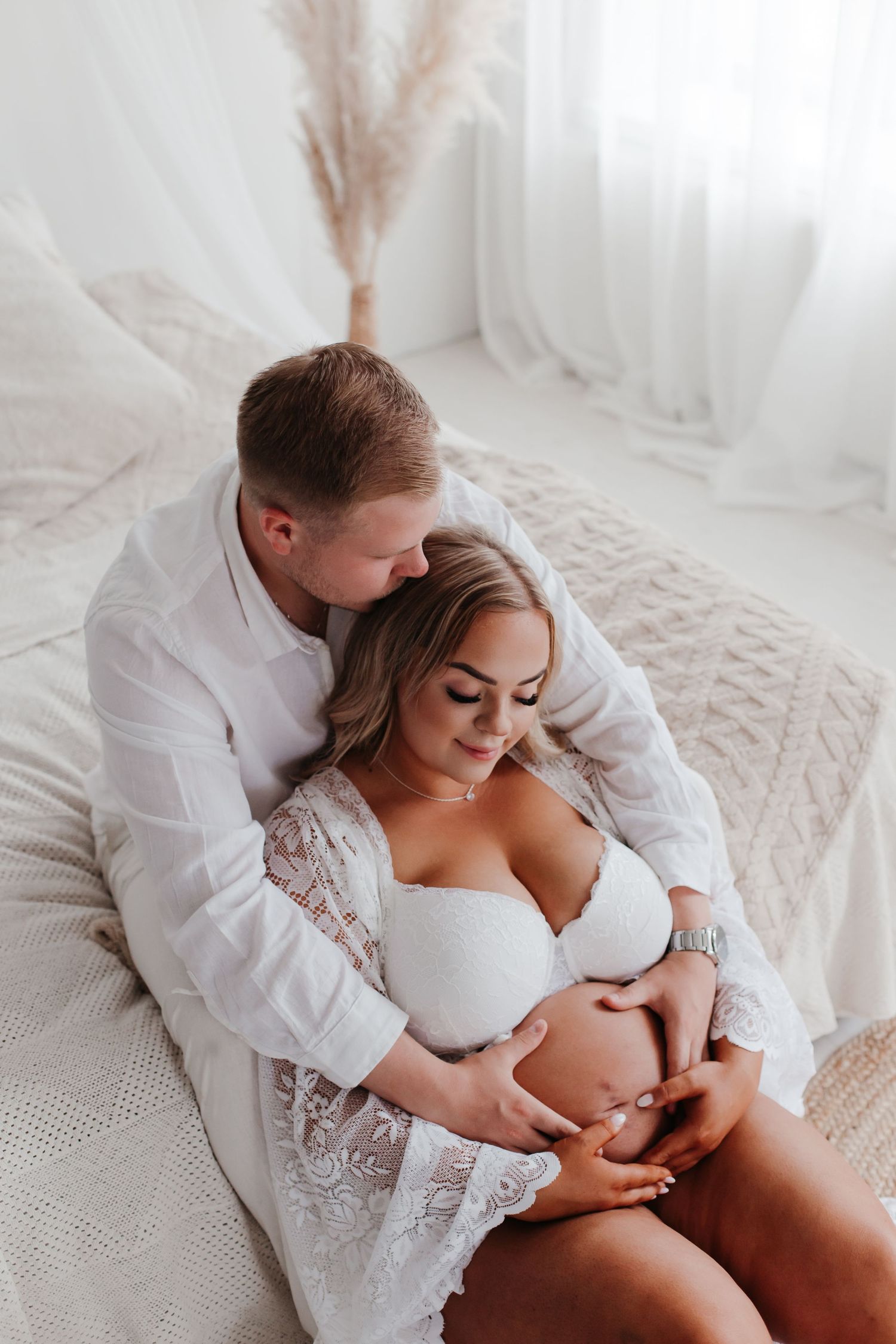 Intimate maternity boudoir photoshoot in white lace lingerie against soft white backdrop.