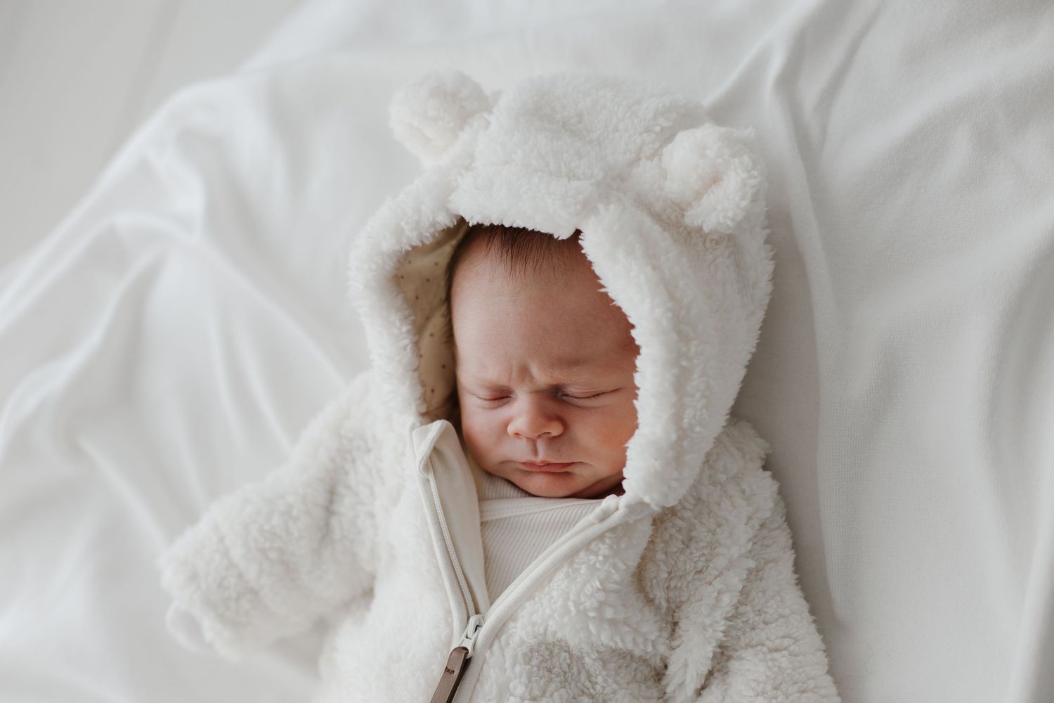 Newborn baby wearing a soft white hooded outfit against light background.