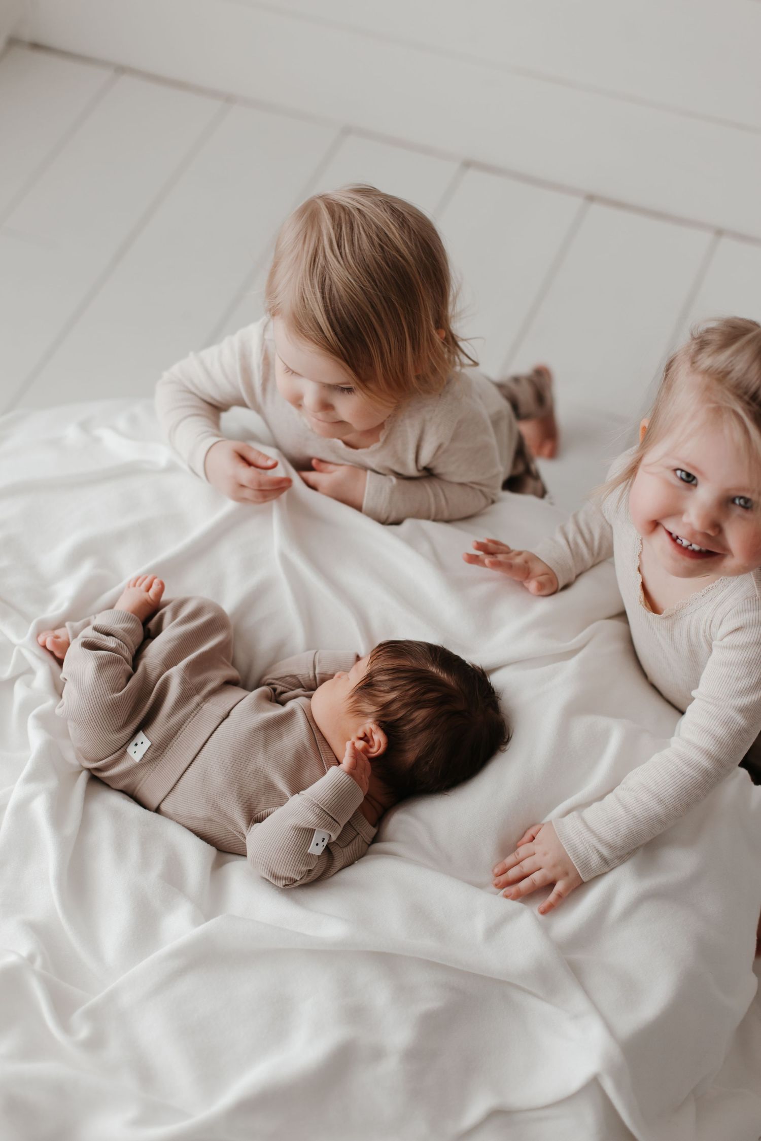 Playful siblings in matching beige outfits sharing joyful moments on white bedding.