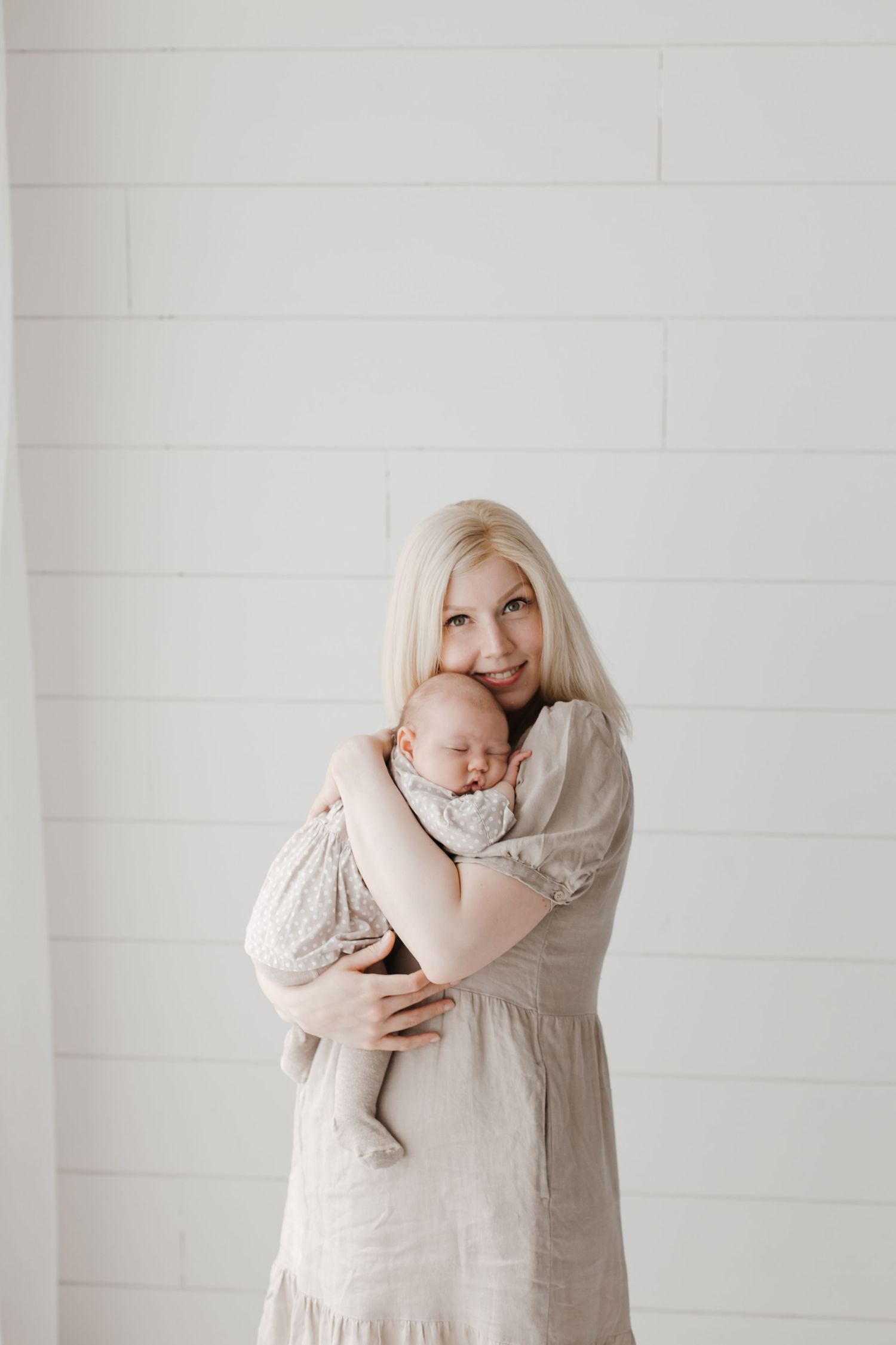 Parent in beige dress holds sleeping infant against white shiplap wall in minimalist setting.