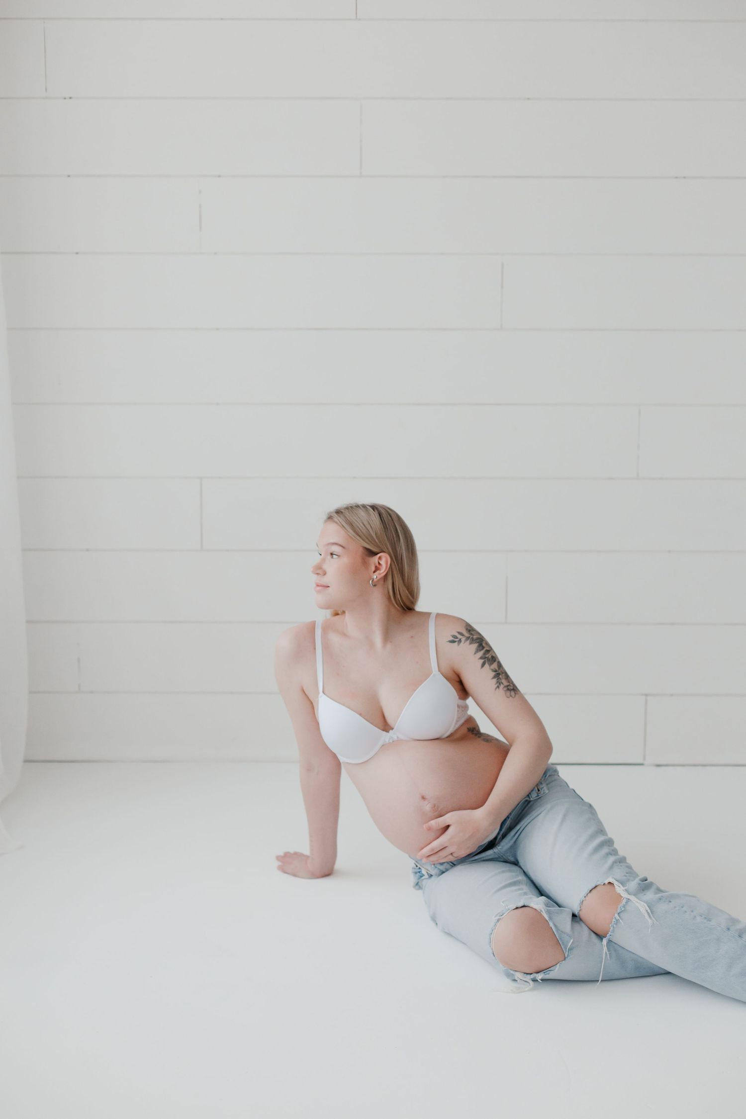 Artistic maternity pose with woman in casual denim and white bra on white floor.