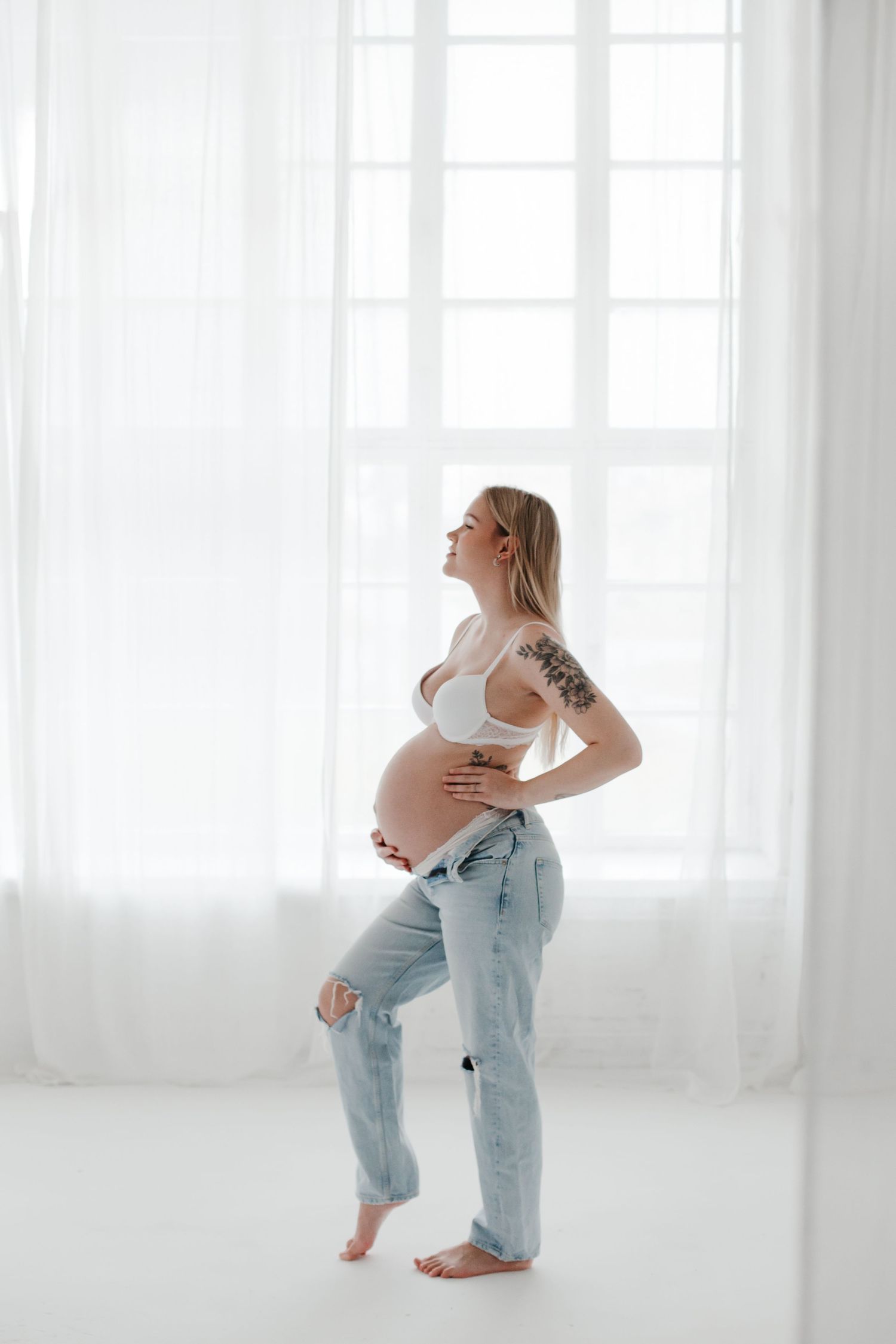 Casual maternity portrait in distressed jeans and white top by bright window.