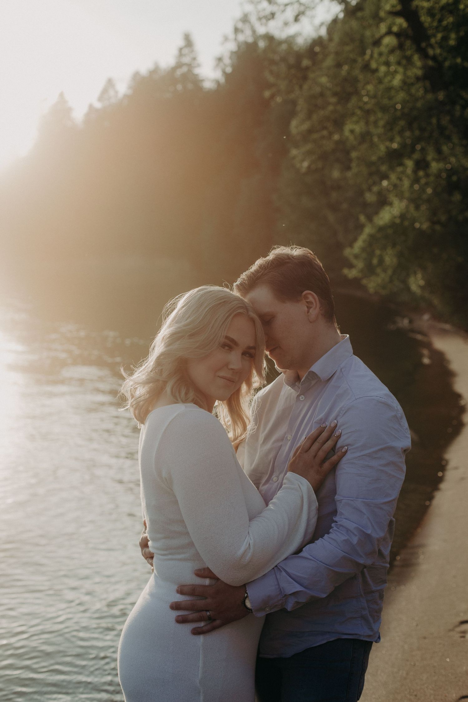 Couple embracing tenderly by a sunlit forest lake shore at golden hour, woman in white dress looking at camera.
