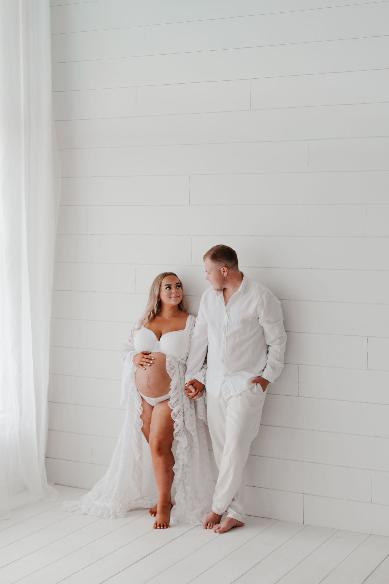 Romantic couple in white attire share tender moment against bright white studio backdrop.