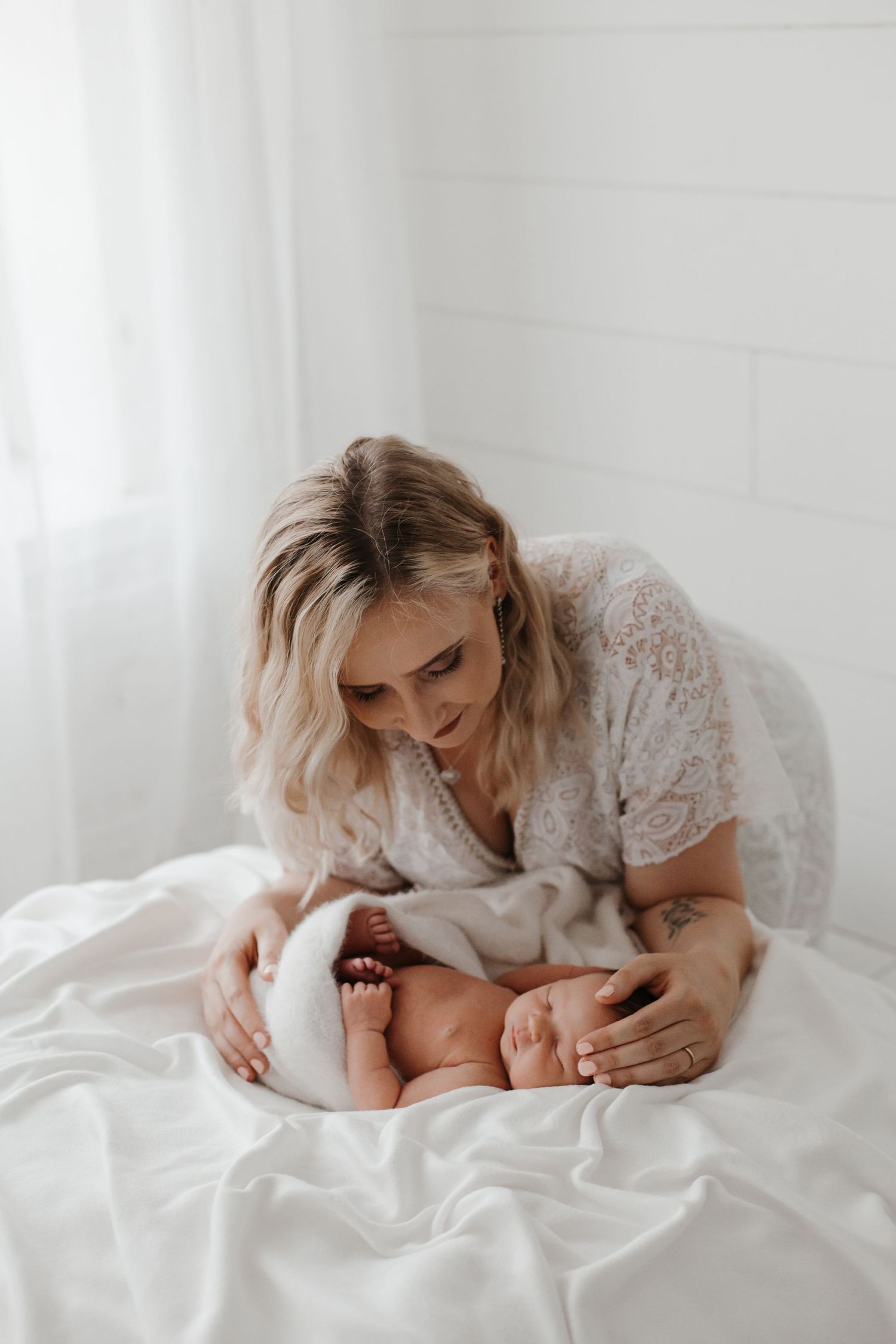 Mother in lace dress cuddling with newborn baby on white bed.