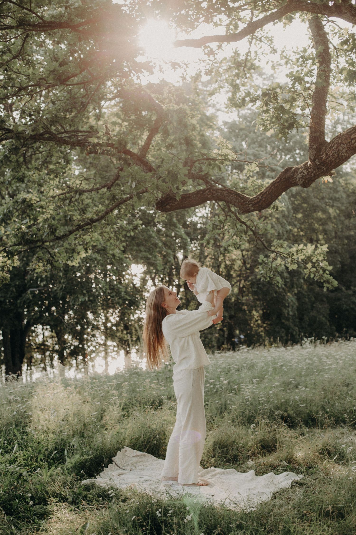 Mother and baby share joyful moment in sunlit outdoor setting with trees.