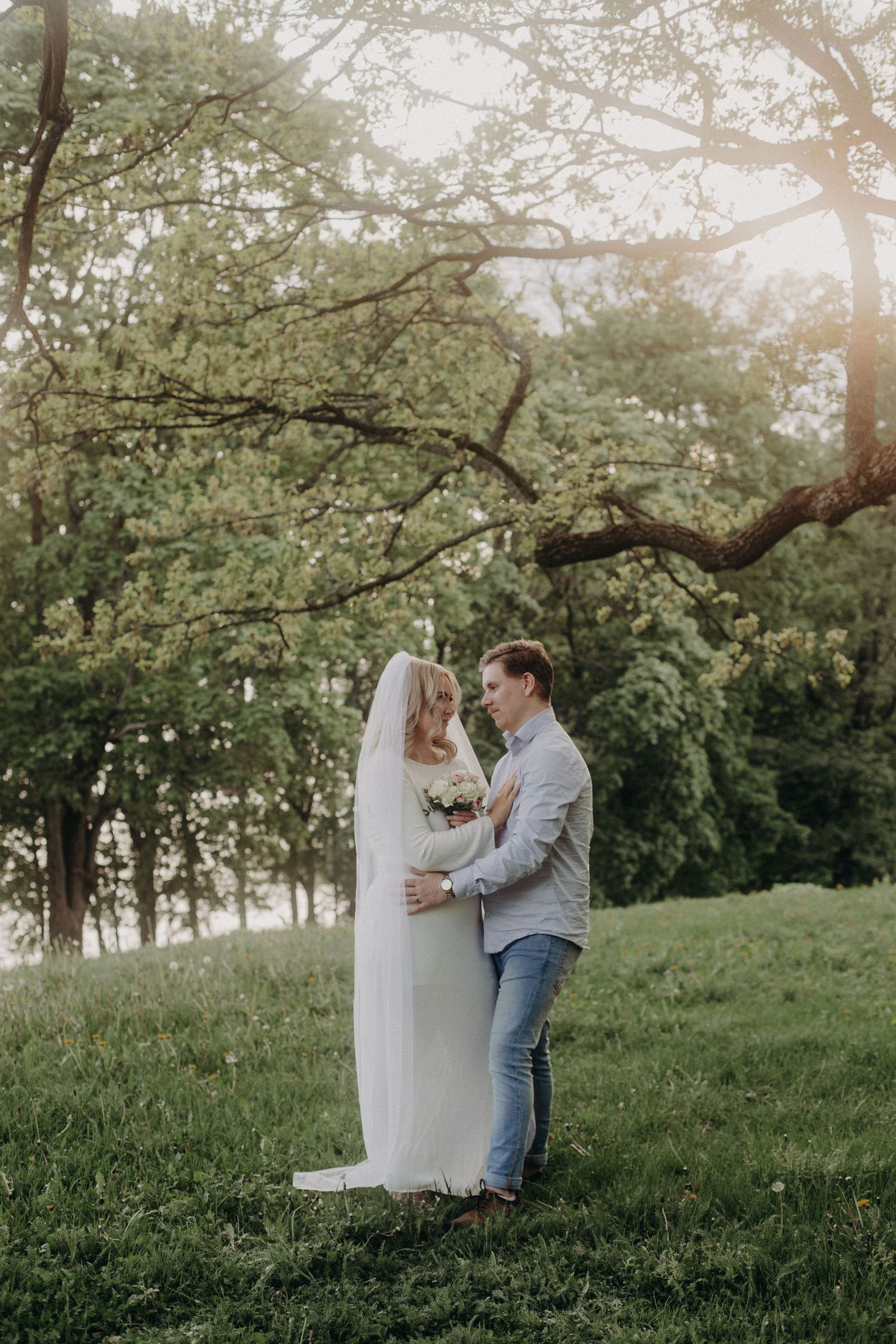 Bride and groom embracing in a lush green park, bride wearing white gown and veil, holding a bouquet at golden hour.