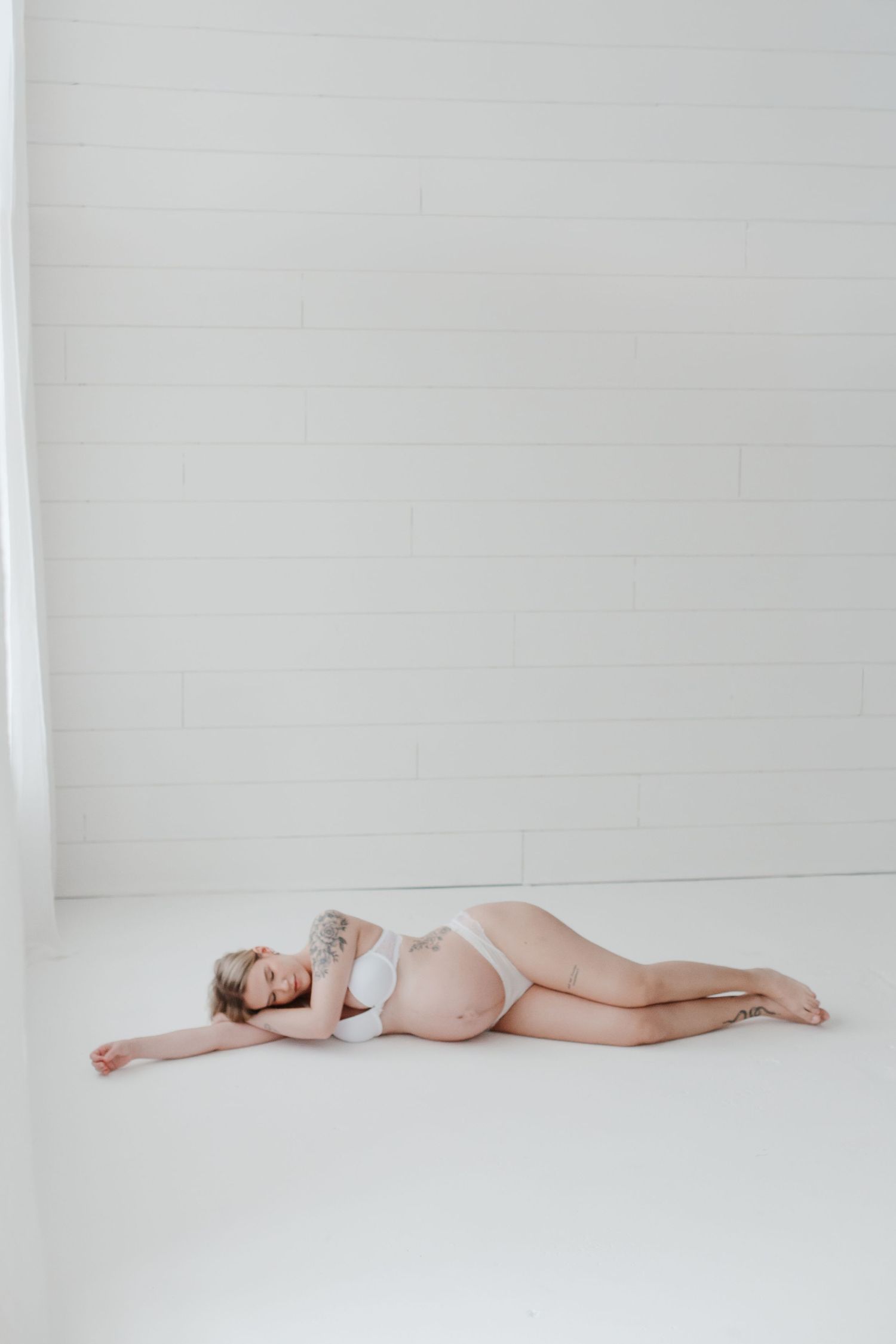 Artistic maternity photo of woman resting on white floor in natural light.