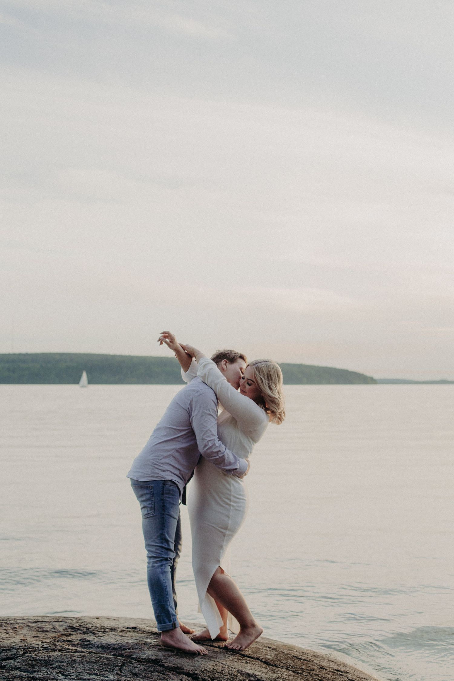 Couple dancing and embracing on rocks by the sea at dusk with a sailboat.