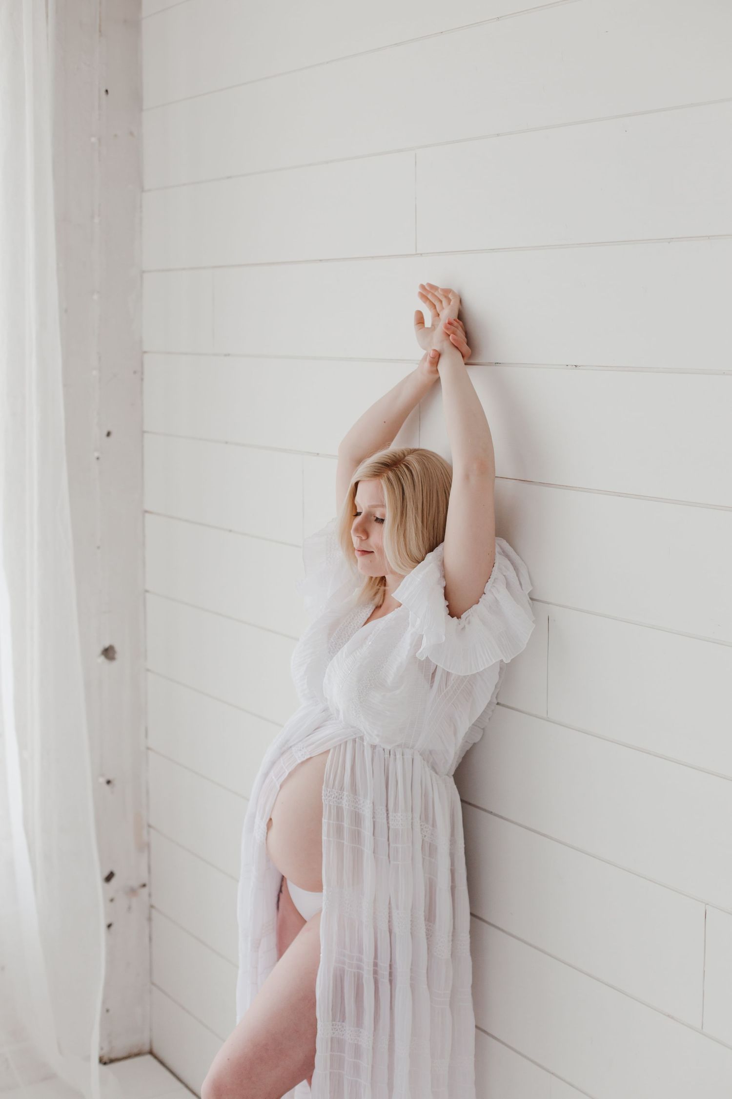 Graceful maternity pose against white wall in sheer white dress.