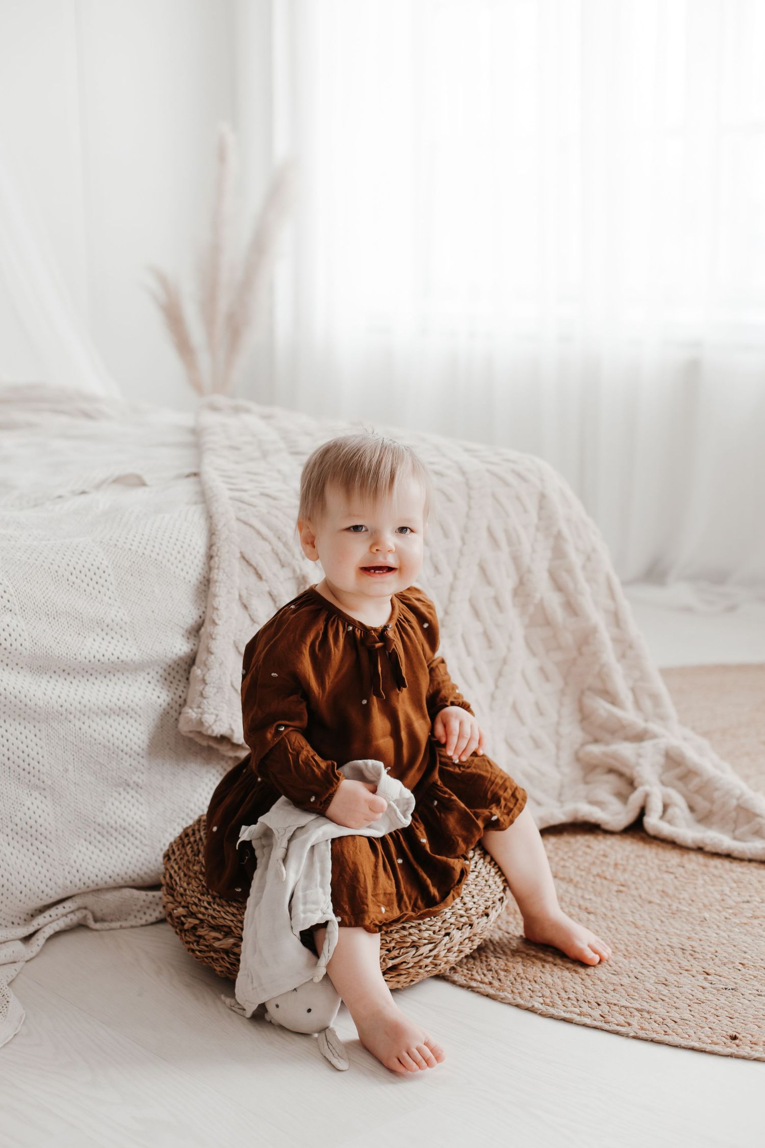 Baby wearing brown dress sits on jute rug in bright white room during portrait session.