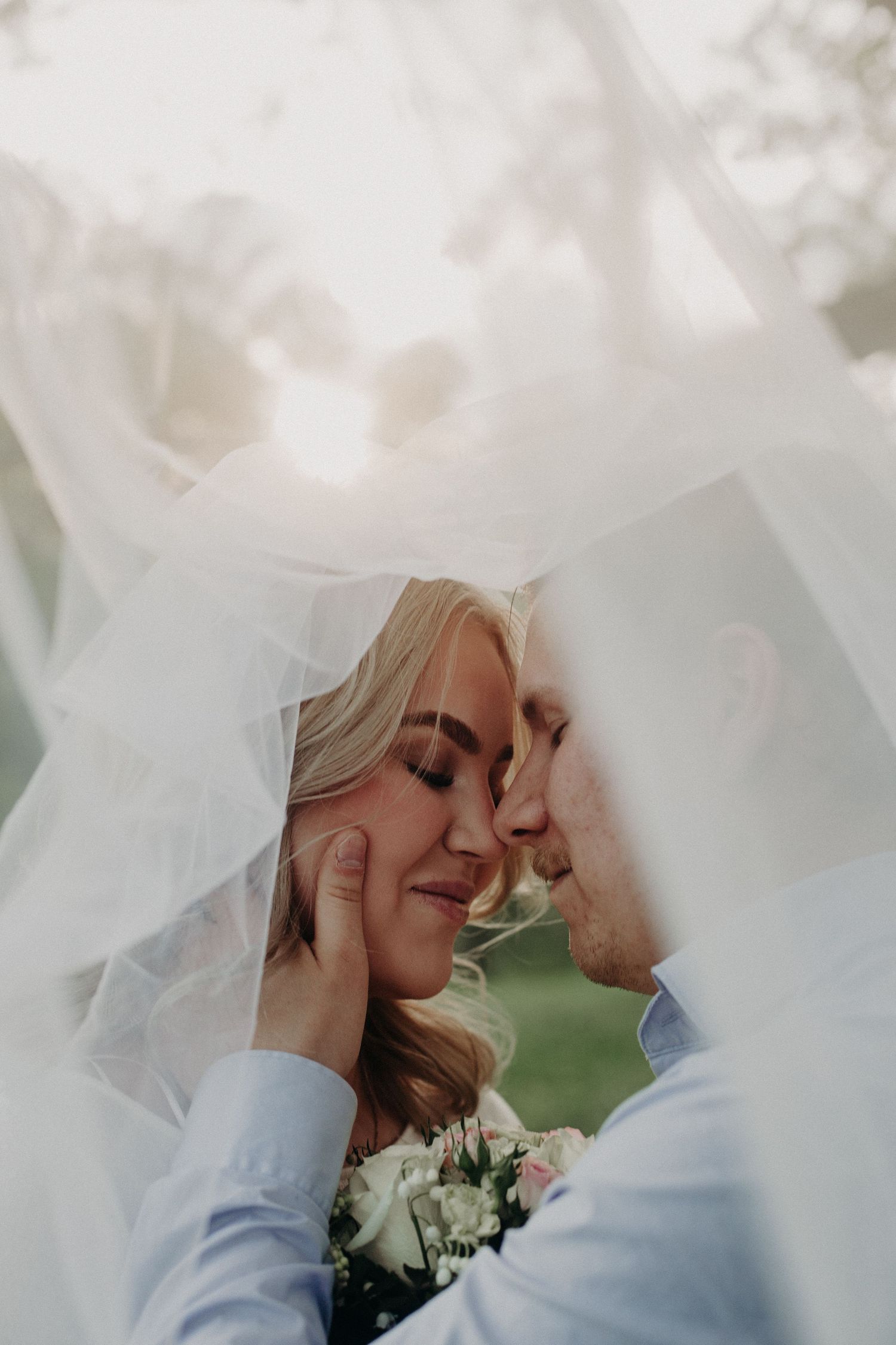Bride and groom sharing a romantic moment under a flowing white veil, with the groom tenderly holding her face.