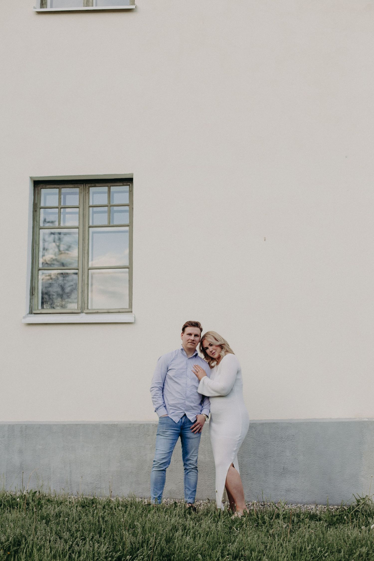 Couple posing together against a white building wall, woman in white dress leaning on man in blue shirt and jeans.