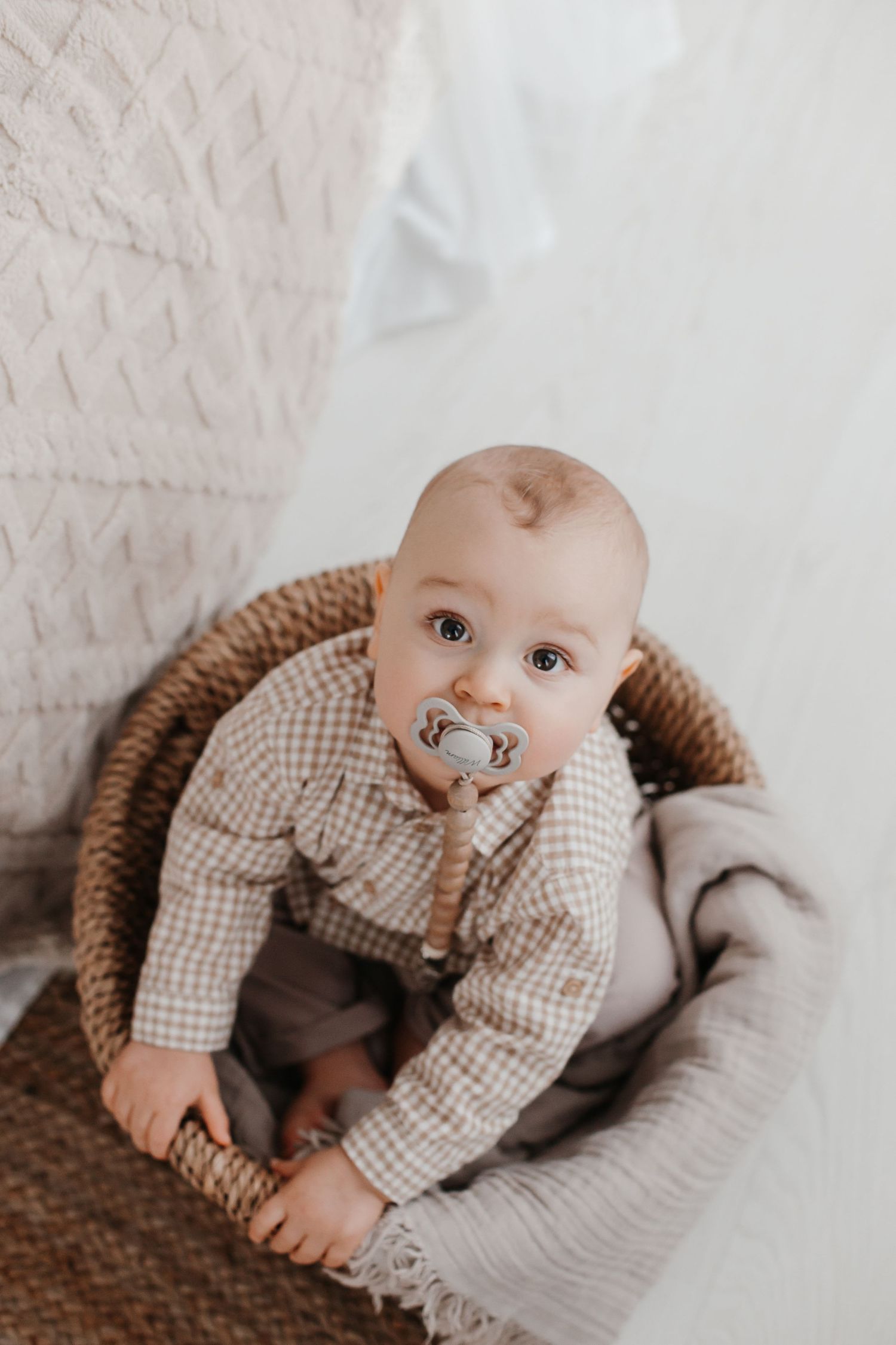 Baby with pacifier wearing checkered shirt sits in woven basket chair.