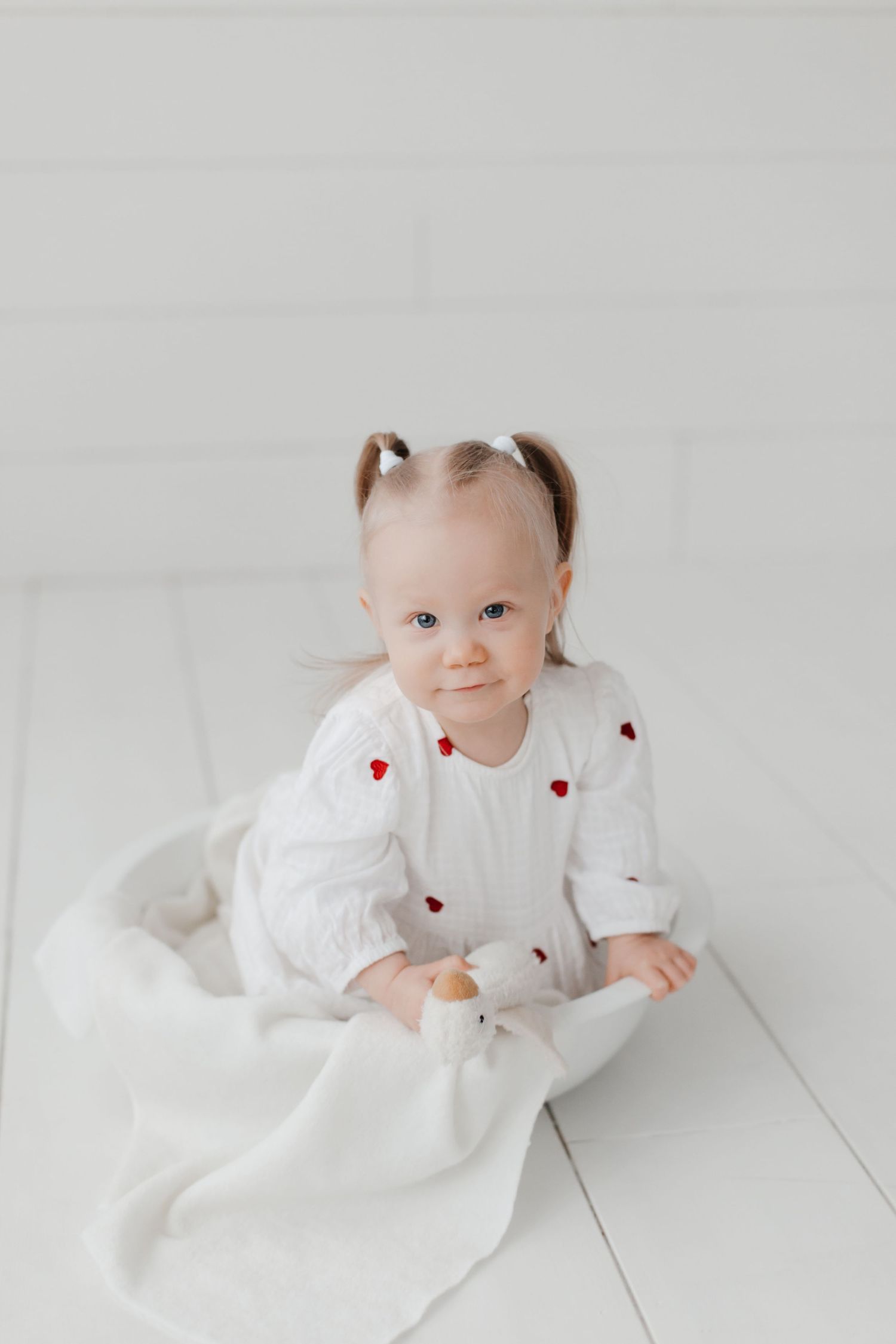 Baby in white heart-patterned dress lying on white floor.