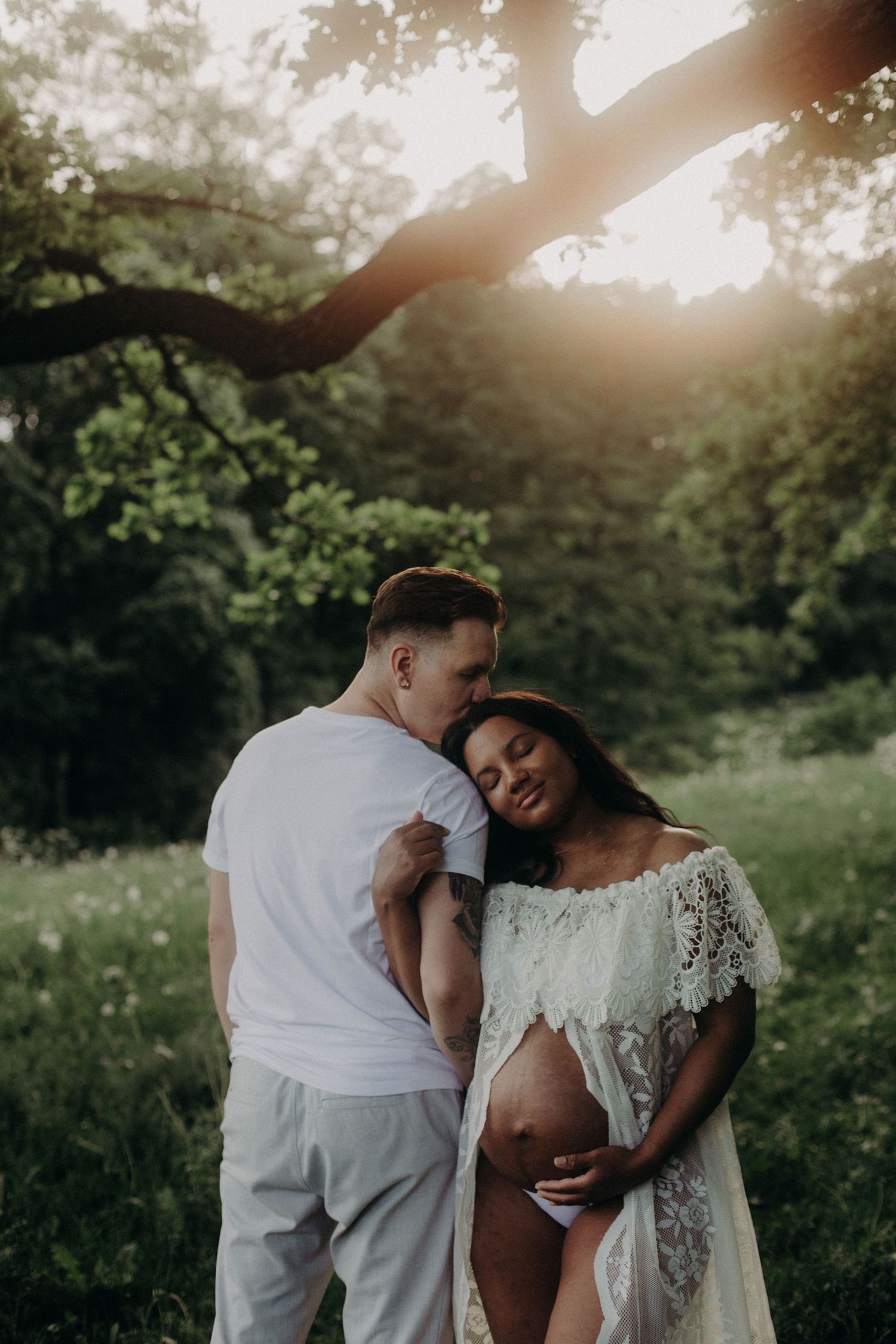 Couple in white sharing a tender maternity moment outdoors at golden hour, pregnant woman in lace dress cradling her bump.