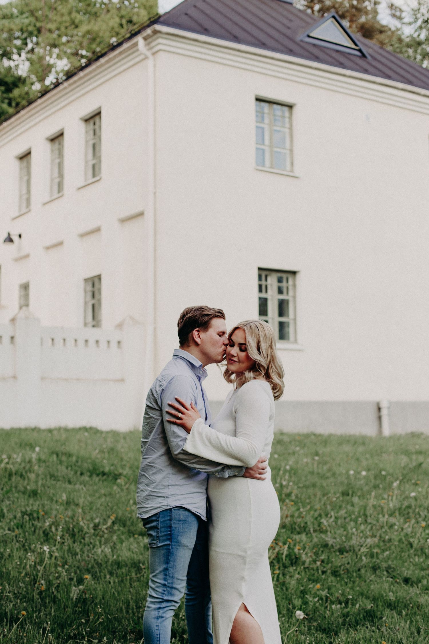 Couple embracing and kissing in front of a white building on a green lawn.