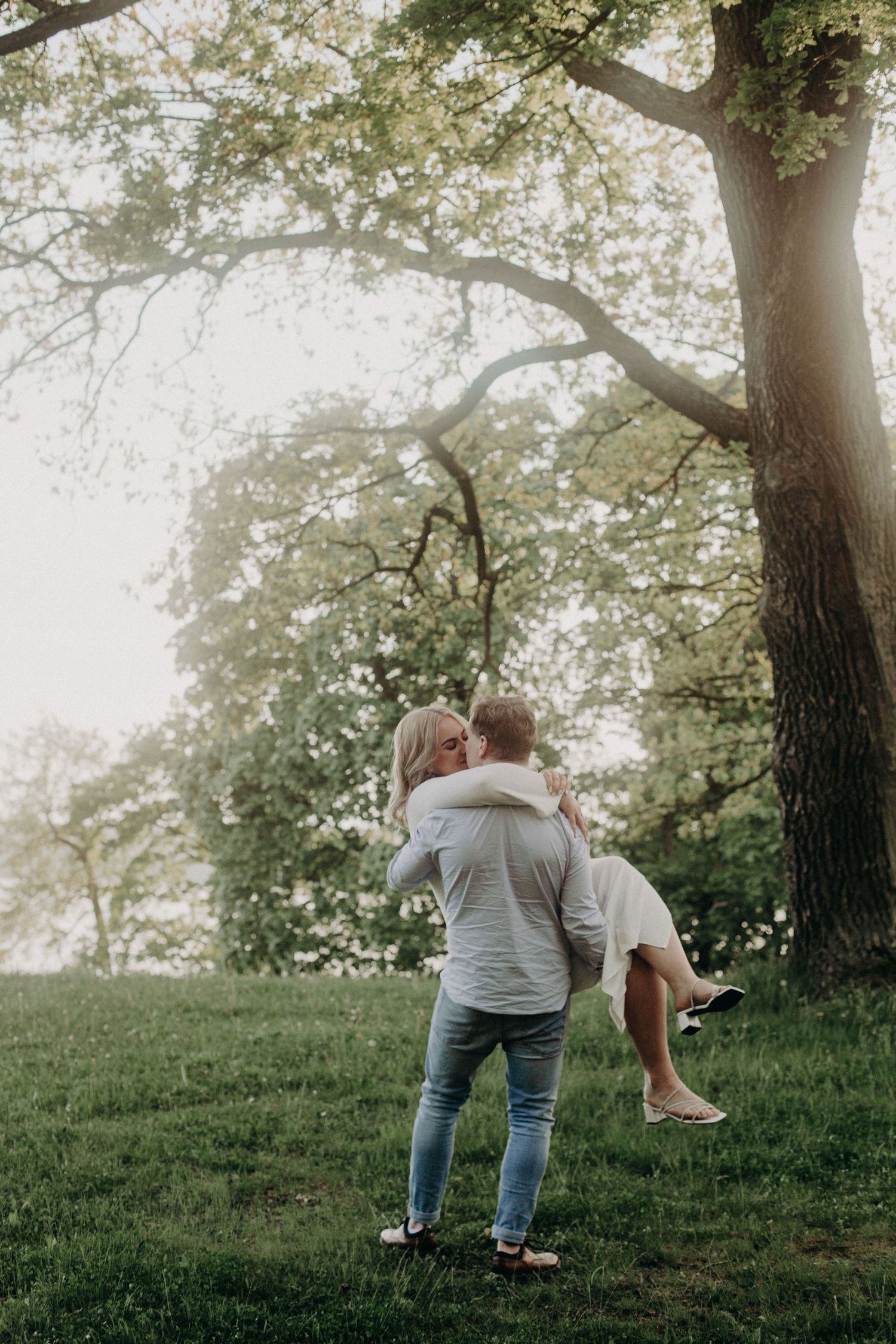 A man lifts a woman in a white dress outdoors under a large tree on a green lawn in soft golden light.